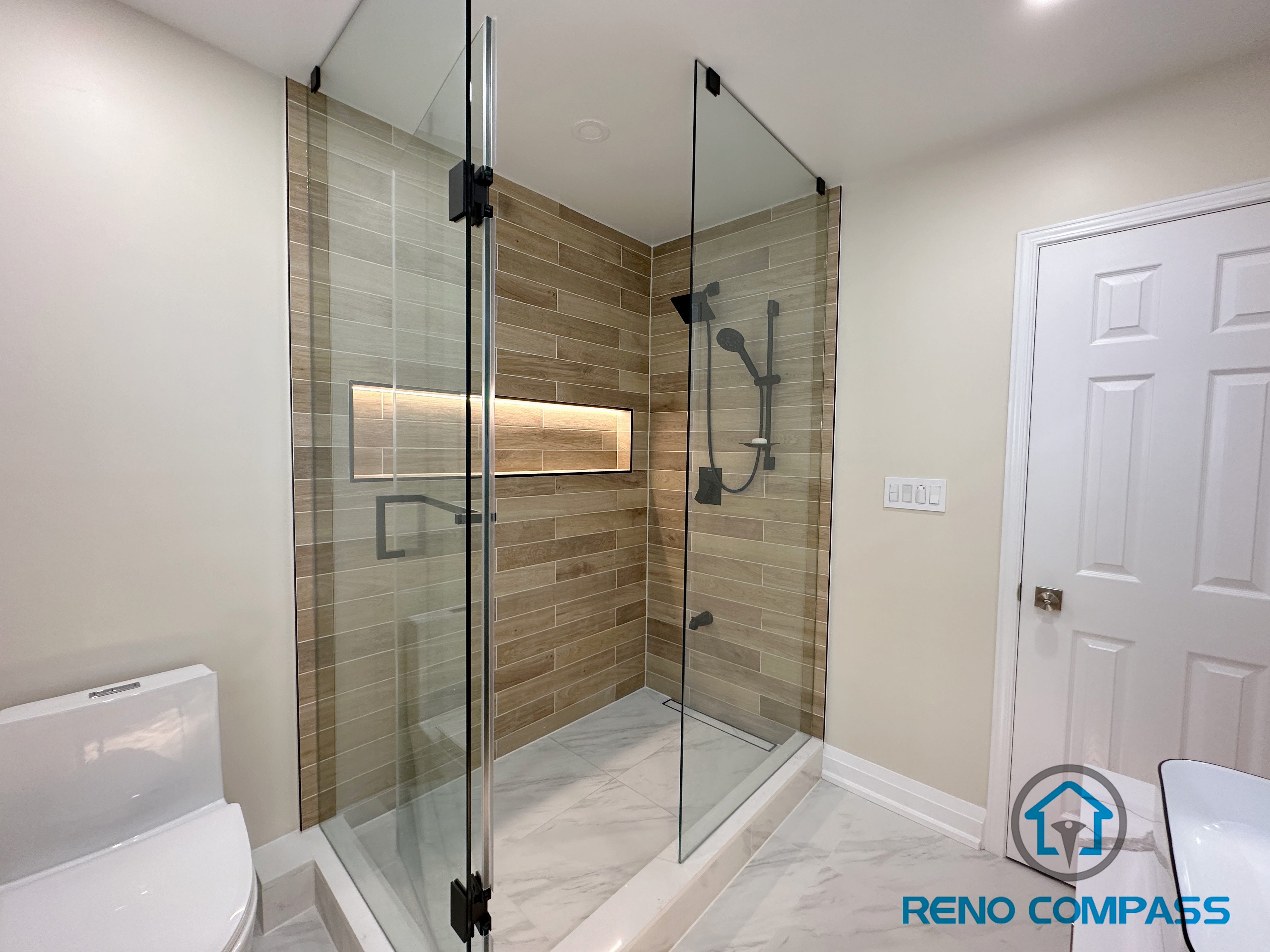 Contemporary bathroom renovation in Vaughan featuring a spacious walk-in shower with a frameless glass enclosure, custom LED-lit shower niche, and wood-look porcelain wall tiles. The shower design includes matte black fixtures, a low-profile shower base, and marble-look porcelain flooring for a seamless and modern finish. This project demonstrates high-quality workmanship and thoughtful layout planning by a trusted Toronto renovation company specializing in custom bathroom renovations.