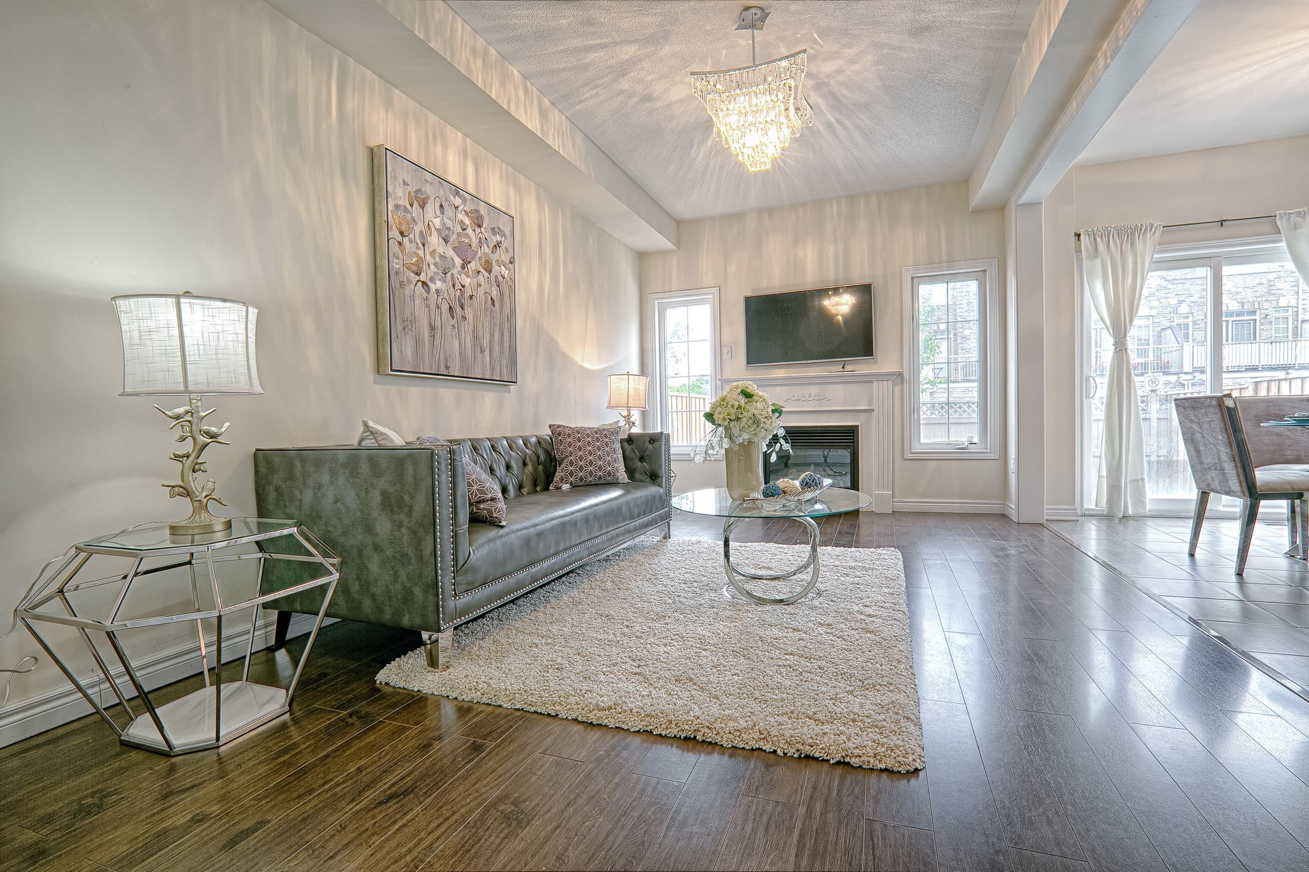 This contemporary chic interior features a refined open-concept living room with dark hardwood flooring, a textured area rug, and a statement crystal chandelier that casts elegant light reflections across the ceiling. The modern fireplace wall with mounted TV, large windows, and soft neutral wall tones create a bright yet cozy atmosphere. Styled with geometric side tables and luxury upholstery, this space reflects the craftsmanship and detail-oriented execution of a trusted Toronto renovation company specializing in modern interior transformations.