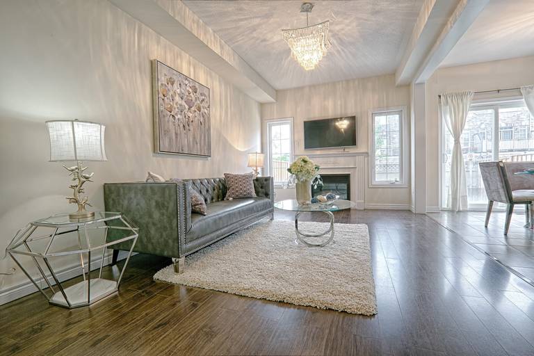 This contemporary chic interior features a refined open-concept living room with dark hardwood flooring, a textured area rug, and a statement crystal chandelier that casts elegant light reflections across the ceiling. The modern fireplace wall with mounted TV, large windows, and soft neutral wall tones create a bright yet cozy atmosphere. Styled with geometric side tables and luxury upholstery, this space reflects the craftsmanship and detail-oriented execution of a trusted Toronto renovation company specializing in modern interior transformations.