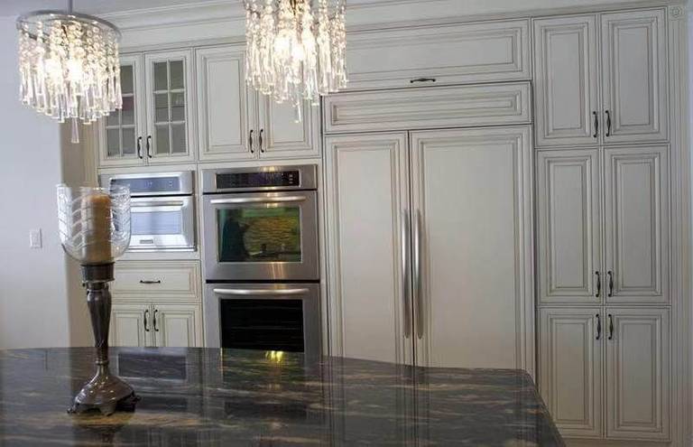 This European-style kitchen renovation showcases elegant custom white cabinetry with detailed panel molding and classic hardware, creating a refined and timeless aesthetic. The kitchen features a fully integrated built-in refrigerator and double wall ovens, maximizing functionality while maintaining a seamless design.

The large central island is finished with a dark natural stone countertop, adding contrast and sophistication to the space. Crystal chandeliers enhance the luxurious European atmosphere, combining traditional charm with modern convenience.

This kitchen design is ideal for homeowners seeking a high-end, classic European kitchen upgrade with enhanced storage, premium finishes, and a cohesive layout.