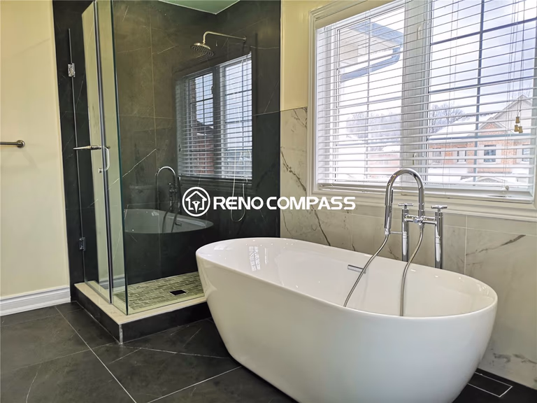 This stylish Toronto bathroom renovation features a sleek freestanding bathtub beside a frameless glass shower. Elegant marble and dark tile flooring create a modern and luxurious spa-like atmosphere. Designed and completed by Reno Compass for a high-end contemporary home upgrade.