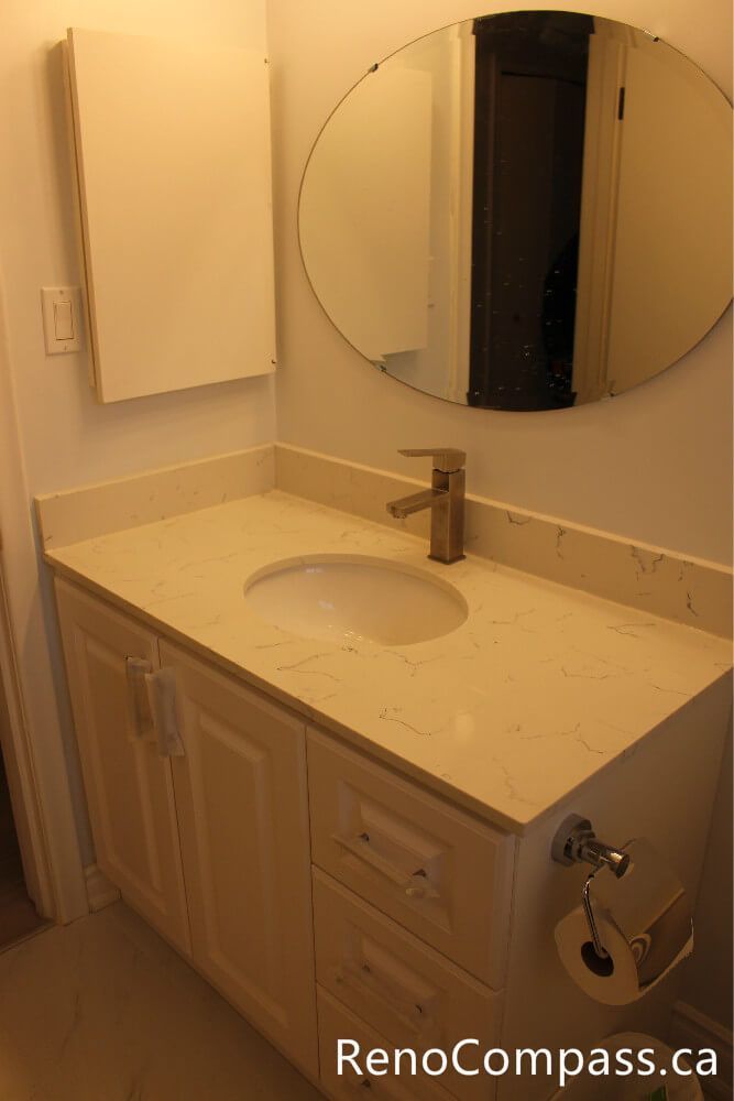 Contemporary bathroom design featuring a white shaker vanity, quartz countertop, and a trendy round wall mirror. Reno Compass specializes in stylish bathroom remodeling in Oshawa, combining functional storage with high-end aesthetic finishes.