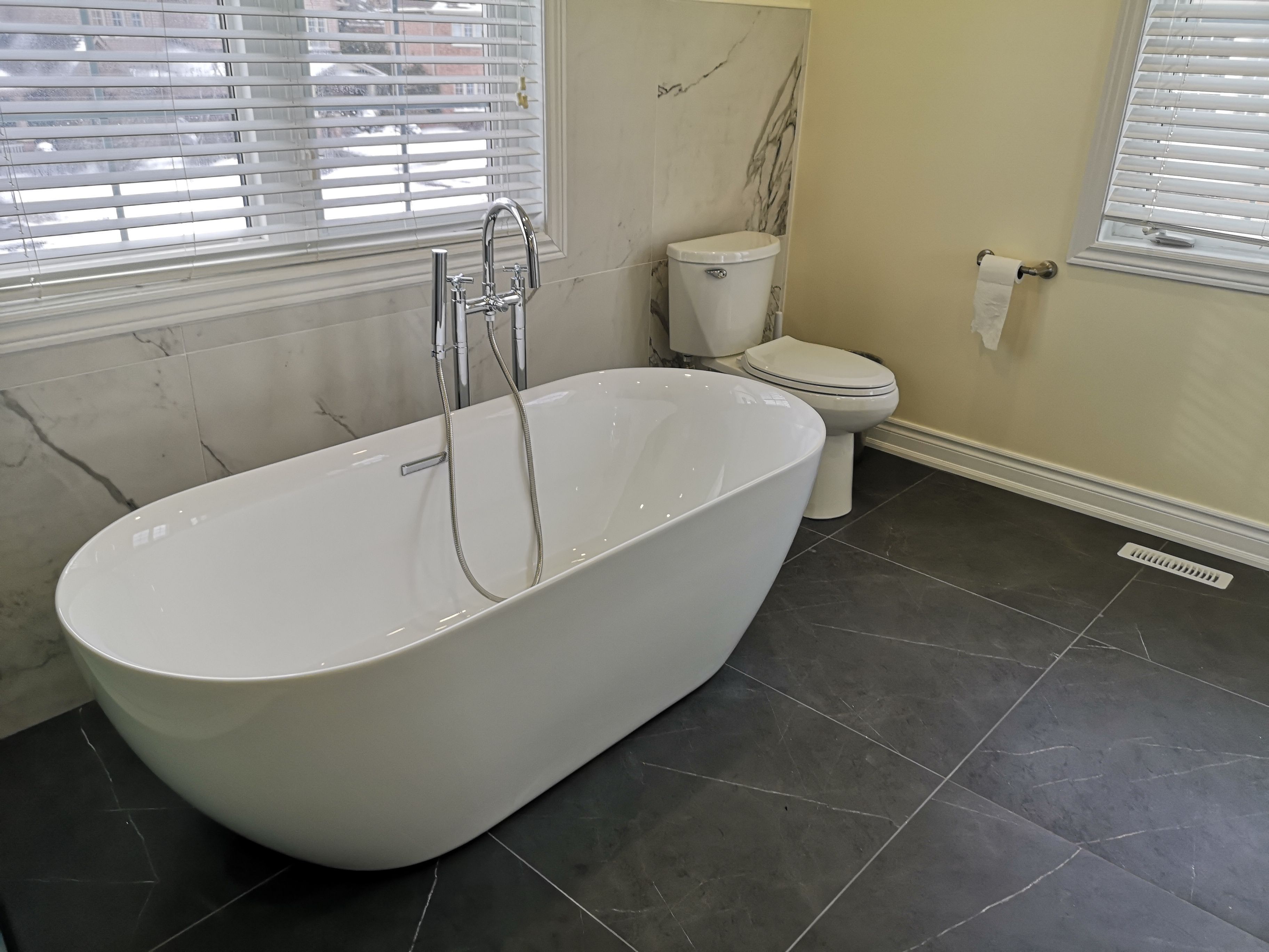 This luxury Toronto bathroom renovation showcases a stunning freestanding bathtub accented by marble wall tiles and sleek dark porcelain flooring. Natural light from dual windows enhances the modern and airy atmosphere, creating a relaxing spa-like retreat. Designed by Reno Compass, the project exemplifies clean lines, balanced contrast, and timeless elegance.