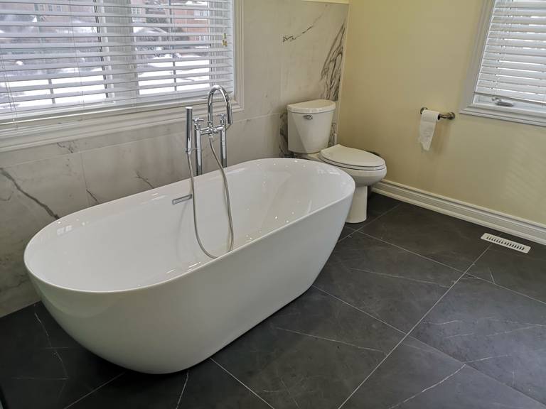 This luxury Toronto bathroom renovation showcases a stunning freestanding bathtub accented by marble wall tiles and sleek dark porcelain flooring. Natural light from dual windows enhances the modern and airy atmosphere, creating a relaxing spa-like retreat. Designed by Reno Compass, the project exemplifies clean lines, balanced contrast, and timeless elegance.