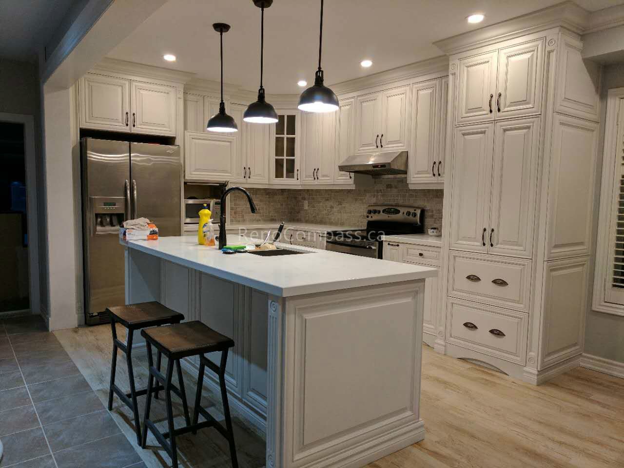 This Vaughan kitchen renovation by Reno Compass Inc. showcases custom white shaker cabinets, a large quartz island with seating, and sleek black pendant lighting. The open-concept layout enhances storage, workflow, and natural light, while stainless steel appliances and warm wood flooring create a clean, modern, and highly functional kitchen designed for daily use and entertaining.
