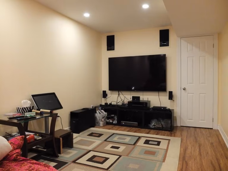 A renovated basement in Vaughan featuring a comfortable home‑theatre area with a wall‑mounted TV, built‑in speakers, entertainment console, warm lighting, and wood‑style flooring. A functional and inviting space designed for relaxation and family entertainment.