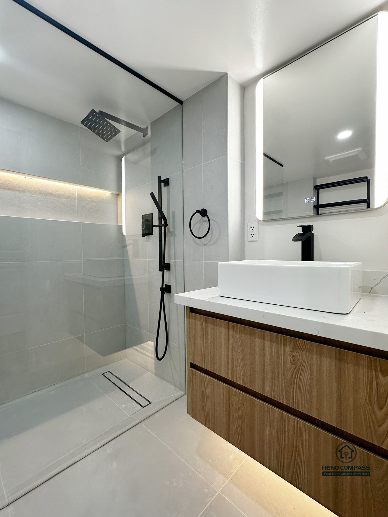 This image captures the relationship between the curbless walk-in shower and the floating vanity, highlighting a well-balanced bathroom layout designed for clarity and ease of use. The seamless shower floor, linear drain, and integrated niche lighting create a clean visual flow, while the warm wood vanity adds contrast and softness to the space. Matte black fixtures and restrained material choices reinforce a modern, understated aesthetic focused on long-term comfort and daily practicality.