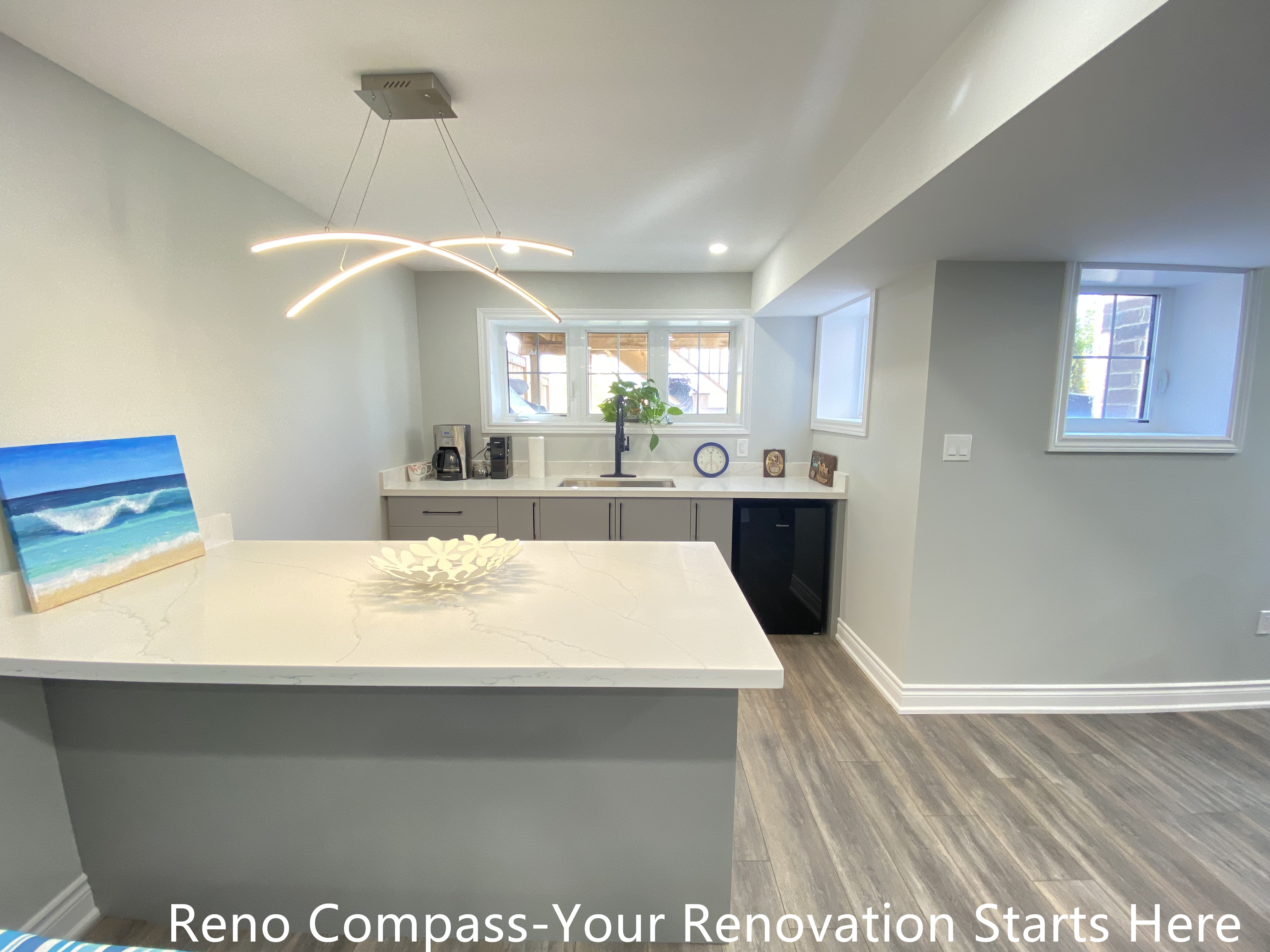 Modern basement bar renovation in Oakville featuring a large quartz island countertop, custom base cabinets, contemporary pendant lighting, vinyl plank flooring, and a compact kitchenette layout designed for entertaining and everyday use, completed by Reno Compass Inc.