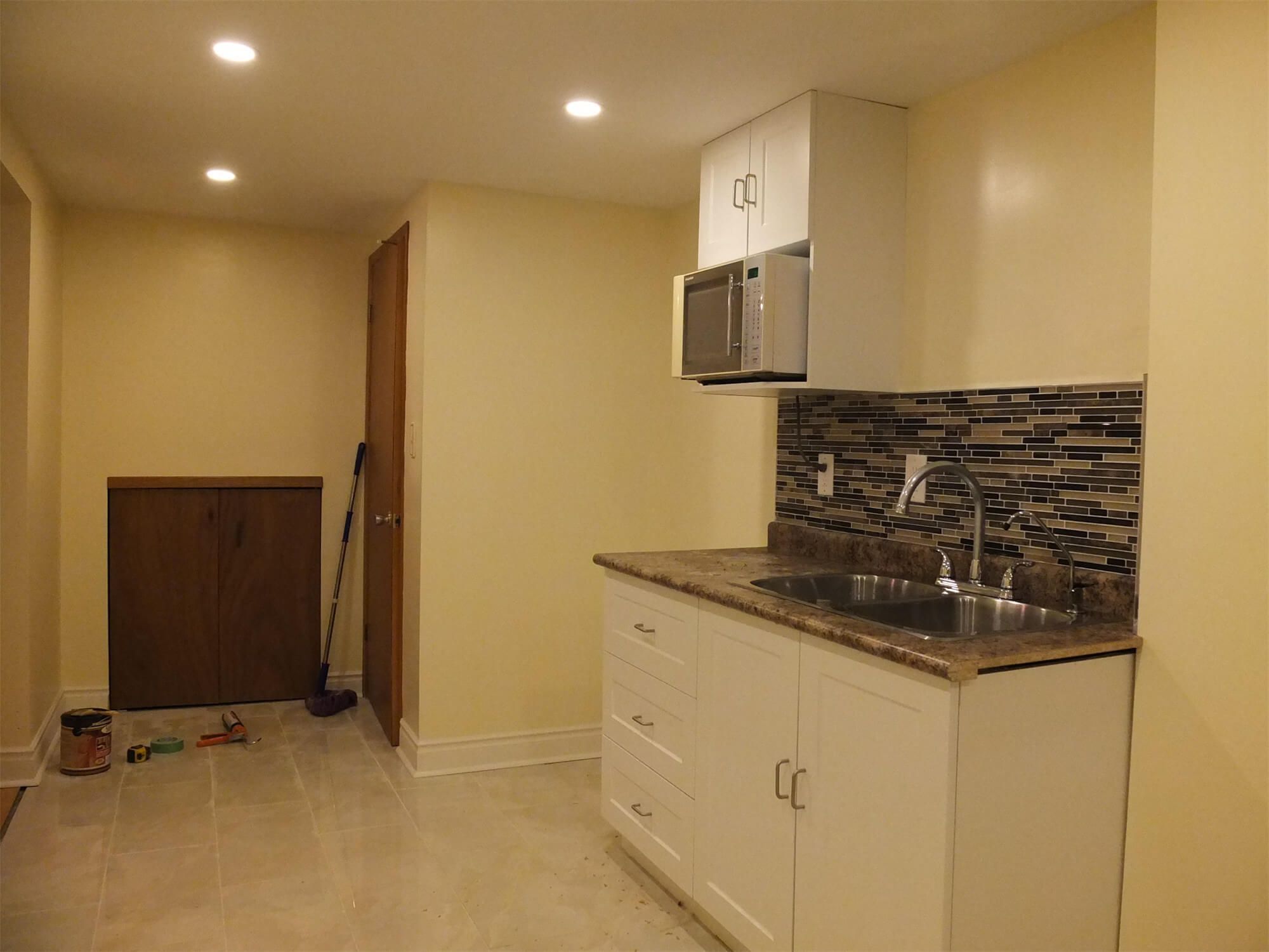 This Oshawa basement renovation features a newly installed kitchenette and laundry area designed for convenience and functionality. The space includes modern white cabinetry, a durable laminate countertop, mosaic tile backsplash, and recessed pot lighting for a clean and practical layout. A stainless-steel sink and built-in microwave enhance usability, while the dedicated washer and dryer setup provides added efficiency for everyday living. The polished tile flooring ensures durability and easy maintenance. Completed by a professional Toronto Renovation Company, this project demonstrates thoughtful planning and quality craftsmanship in transforming a basement into a fully functional living space.