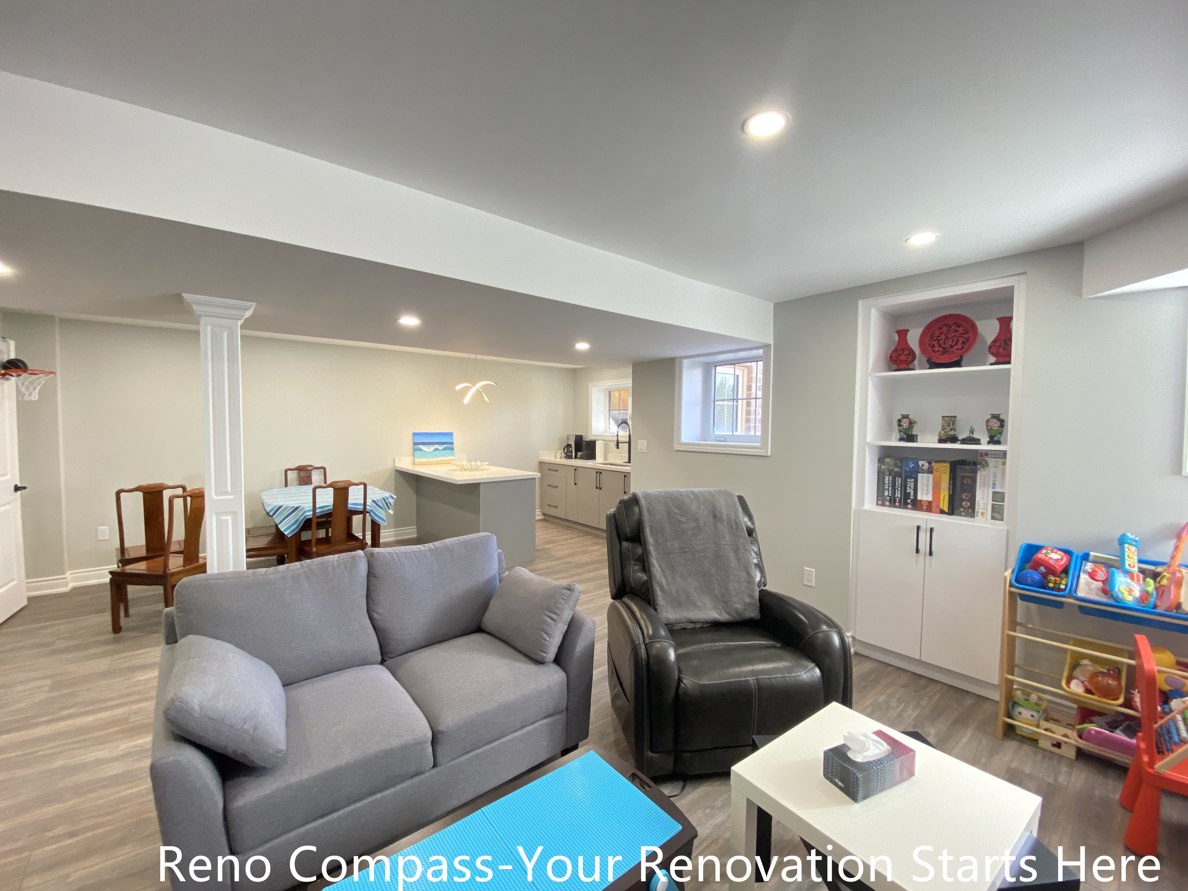 Open-concept basement renovation in Oakville featuring a spacious family living area with recessed lighting, structural columns, vinyl plank flooring, built-in shelving, and seamless connection to a custom bar area, creating a bright and functional multi-purpose basement space, completed by Reno Compass Inc.