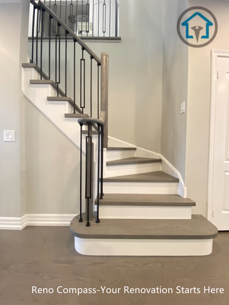This custom staircase renovation features a gently curved entry step, warm wood treads, and slim black metal balusters that bring structure and elegance to the space. The curved base softens the transition between floors while improving circulation and visual balance. Clean white risers and consistent detailing create a refined, cohesive look that integrates seamlessly with the surrounding interior finishes, completed as part of a full-home renovation by Reno Compass.