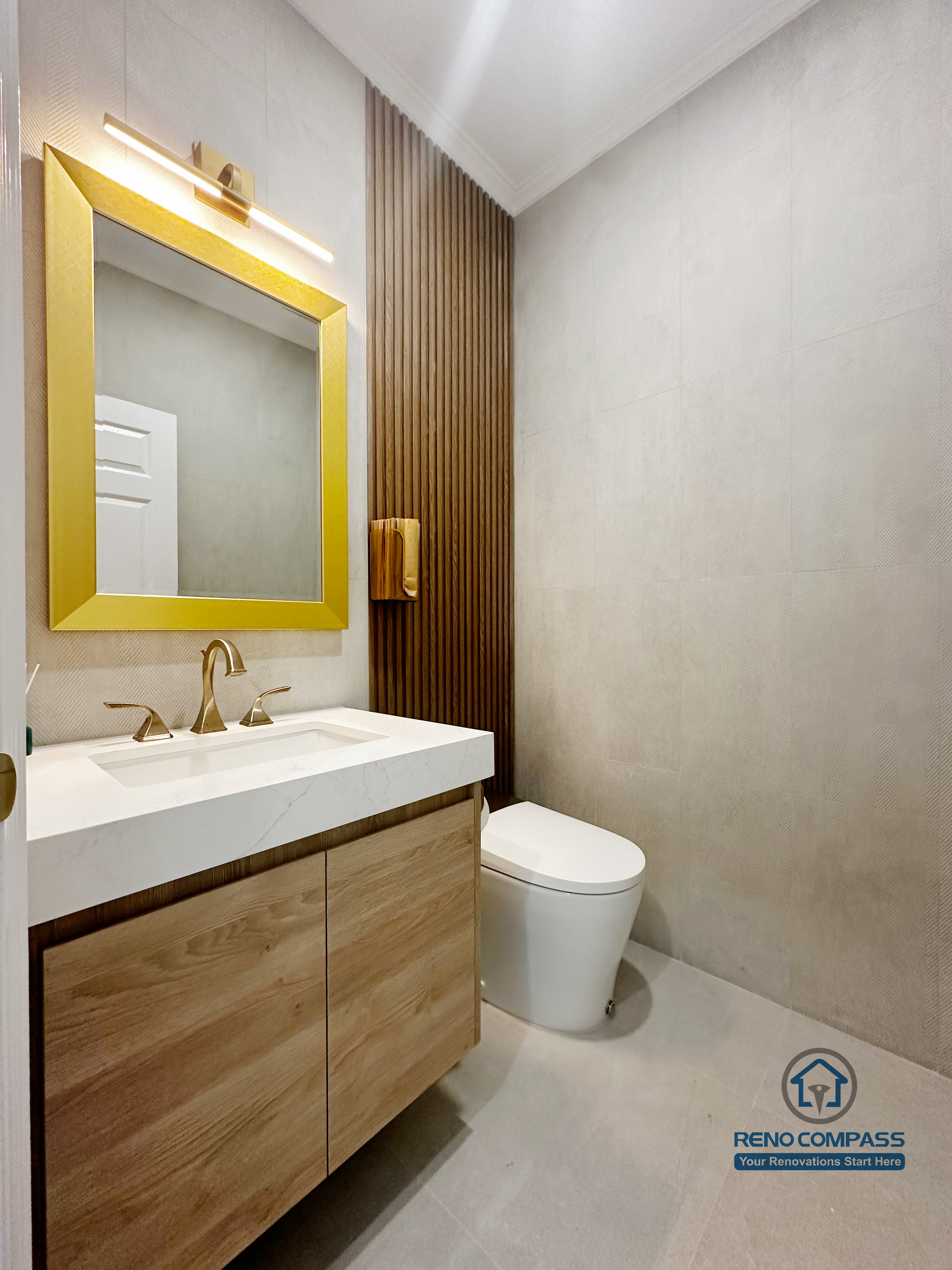This bathroom renovation image highlights a modern powder room design in North York, featuring a custom wood vanity paired with a white integrated sink and brushed gold faucet. The bold gold-framed mirror and matching wall sconce add a refined focal point, while the vertical wood slat accent behind the toilet introduces warmth and visual rhythm. Large-format neutral tiles and clean lines create a balanced, contemporary bathroom layout with strong attention to detail and material coordination.