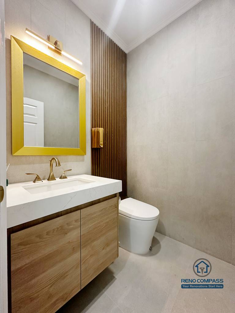 This bathroom renovation image highlights a modern powder room design in North York, featuring a custom wood vanity paired with a white integrated sink and brushed gold faucet. The bold gold-framed mirror and matching wall sconce add a refined focal point, while the vertical wood slat accent behind the toilet introduces warmth and visual rhythm. Large-format neutral tiles and clean lines create a balanced, contemporary bathroom layout with strong attention to detail and material coordination.