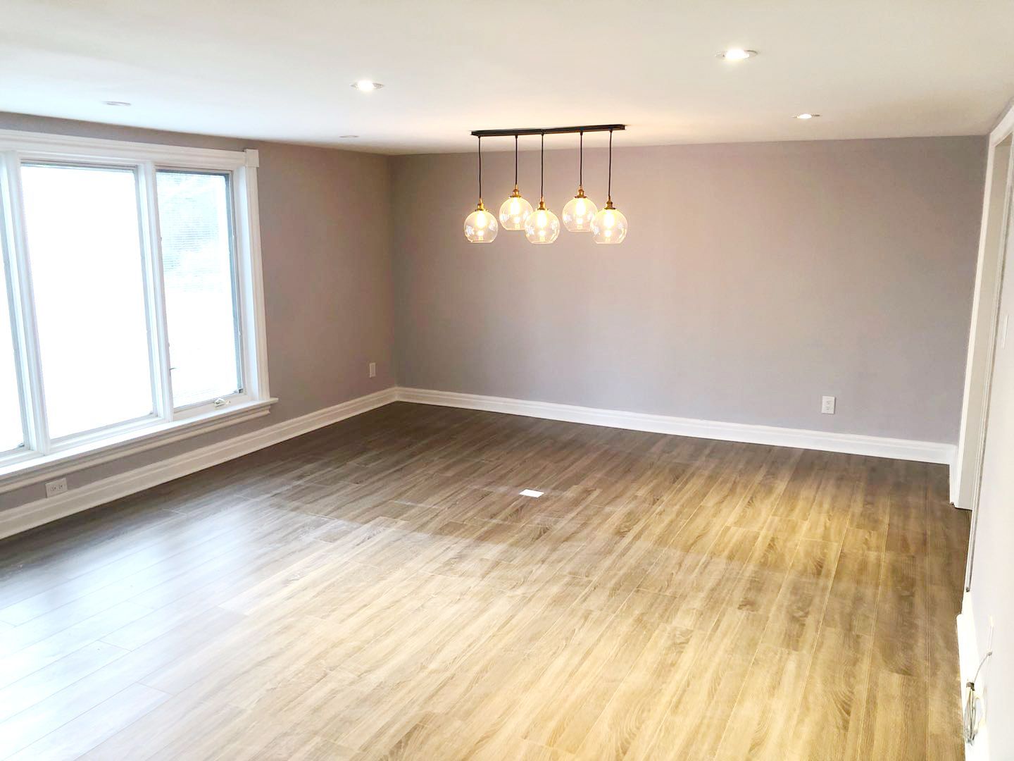 This Etobicoke interior renovation showcases a spacious open living area with upgraded flooring, neutral wall tones, and modern pendant lighting that creates a warm and inviting atmosphere. Large windows allow natural light to brighten the space, while recessed ceiling lights enhance overall illumination. Designed and completed by Reno Compass Inc., this renovation focuses on clean finishes, practical layout, and timeless interior design that improves everyday comfort and functionality.