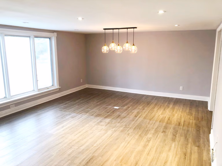 This Etobicoke interior renovation showcases a spacious open living area with upgraded flooring, neutral wall tones, and modern pendant lighting that creates a warm and inviting atmosphere. Large windows allow natural light to brighten the space, while recessed ceiling lights enhance overall illumination. Designed and completed by Reno Compass Inc., this renovation focuses on clean finishes, practical layout, and timeless interior design that improves everyday comfort and functionality.