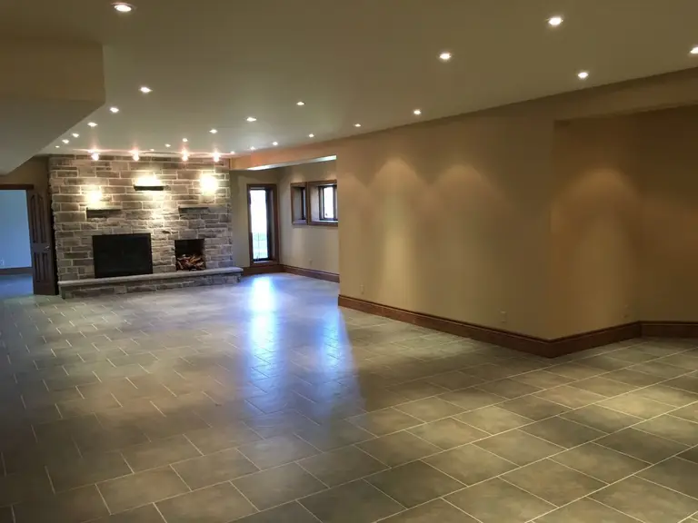 This Toronto interior renovation showcases a spacious open-concept living area designed with modern large-format tile flooring, warm neutral wall tones, and elegant recessed lighting throughout the ceiling. A beautifully crafted stone fireplace feature wall creates a strong focal point, adding both warmth and architectural character to the space. The upgraded lighting layout enhances brightness and visual depth, making the area ideal for entertaining and everyday living. Completed by a professional Toronto renovation company, this project highlights quality finishes, clean design lines, and a refined contemporary interior transformation.