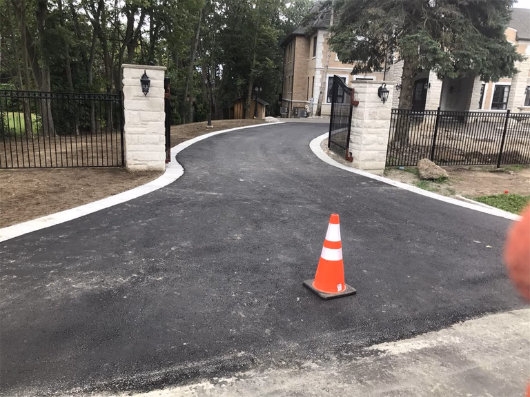 Elegant Toronto driveway landscaping project featuring newly paved asphalt driveway, curved entrance layout, decorative stone gate pillars, and black metal fencing. The design creates a welcoming residential entrance while improving driveway functionality and curb appeal.