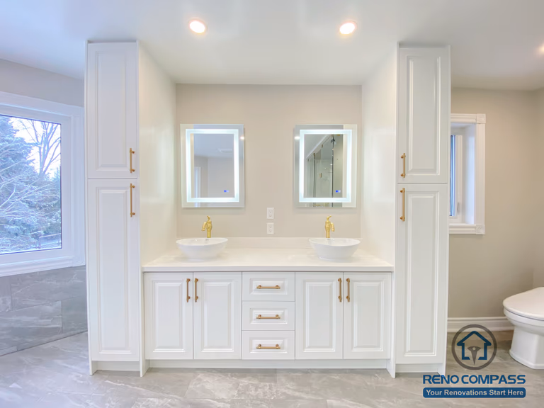 This Richmond Hill bathroom renovation features a custom-built double vanity with integrated storage cabinets and symmetrical design. The white cabinetry is paired with gold hardware, vessel sinks, and backlit mirrors, creating a clean and elegant focal point. Tall storage towers on both sides enhance functionality while maintaining visual balance. This vanity area reflects thoughtful space planning, refined material selection, and high-quality craftsmanship in a modern bathroom renovation project in Richmond Hill.