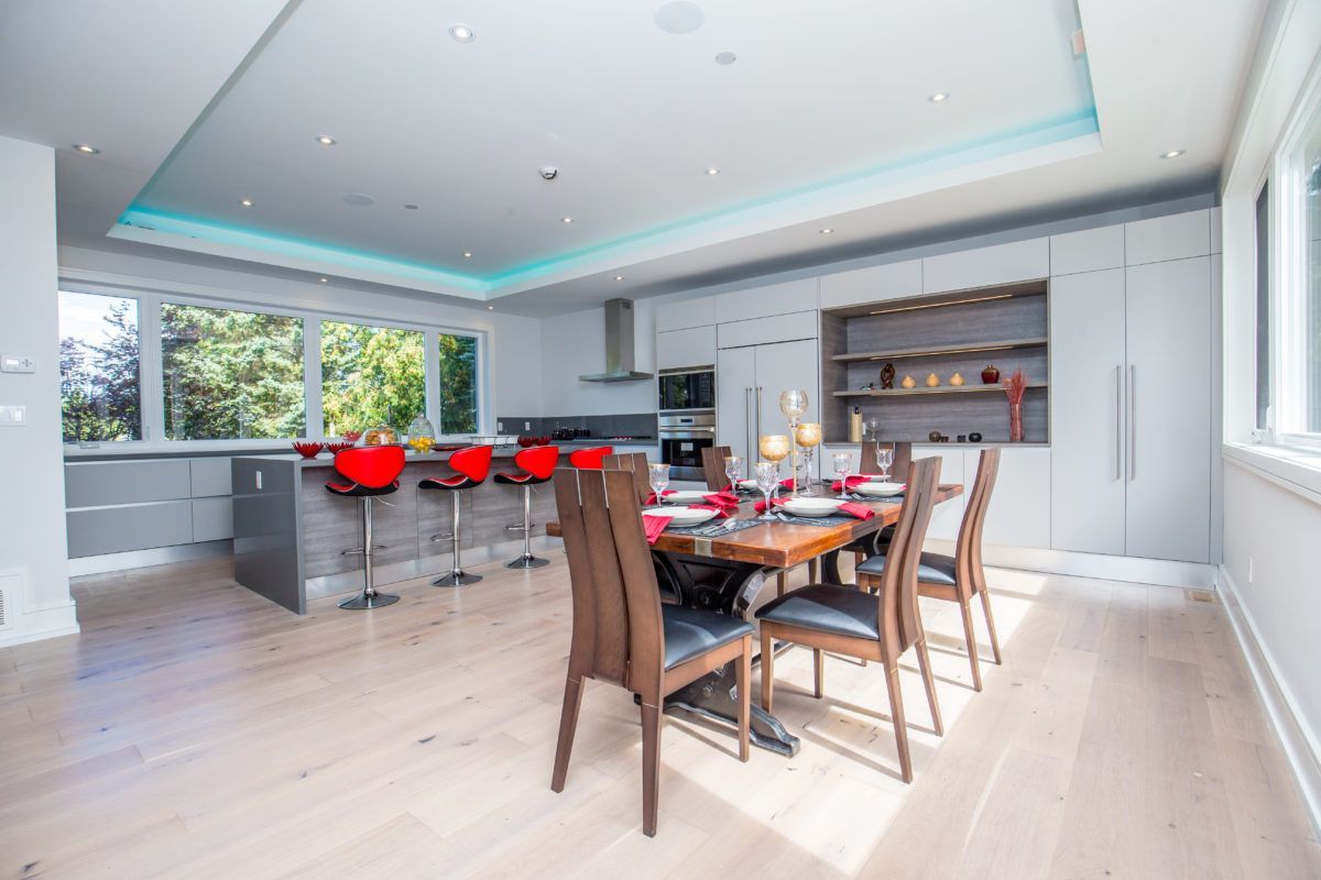 Contemporary open-concept kitchen and dining area in a 5000 sqft Toronto interior renovation, featuring custom grey cabinetry, a large kitchen island with red bar stools, built-in stainless steel appliances, and wide plank hardwood flooring. The dining space is styled with a solid wood table and modern seating, creating a seamless flow between cooking and entertaining areas. Completed by Reno Compass Inc., a trusted Toronto renovation company specializing in luxury home interior remodeling and large-scale residential upgrades.
