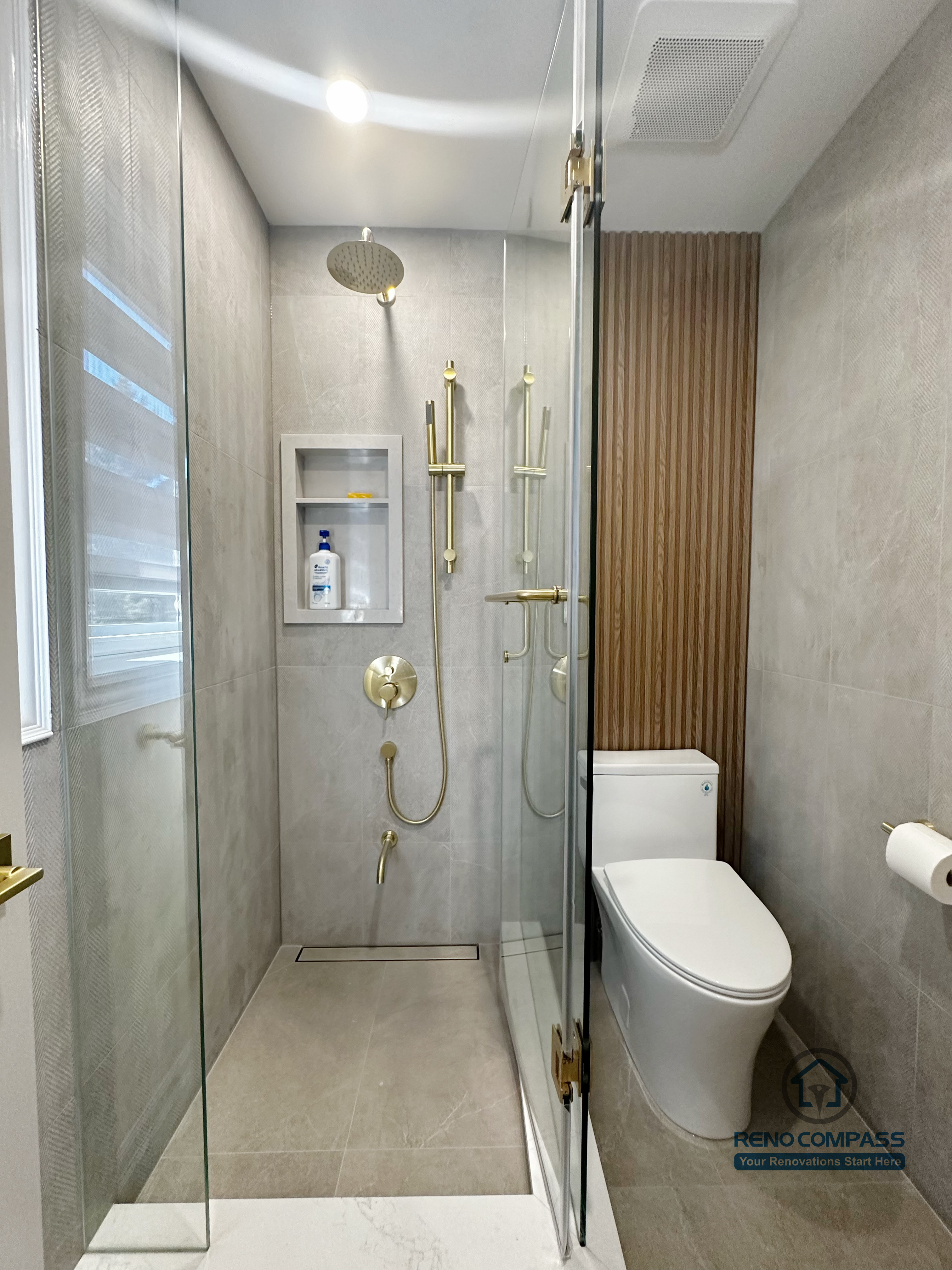 This image highlights a walk-in shower from a North York bathroom renovation, featuring a full-height glass enclosure and large-format grey wall tiles. A built-in shower niche provides practical storage while maintaining a clean, seamless surface. The gold rain shower system and matching controls add warmth and contrast to the neutral palette, while the vertical wood slat accent beside the toilet introduces texture and architectural detail. This layout balances modern aesthetics with efficient use of space and everyday functionality.