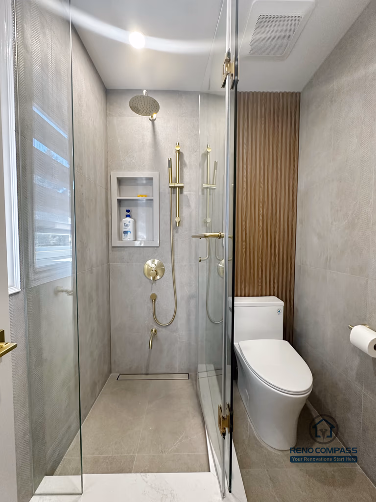 This image highlights a walk-in shower from a North York bathroom renovation, featuring a full-height glass enclosure and large-format grey wall tiles. A built-in shower niche provides practical storage while maintaining a clean, seamless surface. The gold rain shower system and matching controls add warmth and contrast to the neutral palette, while the vertical wood slat accent beside the toilet introduces texture and architectural detail. This layout balances modern aesthetics with efficient use of space and everyday functionality.