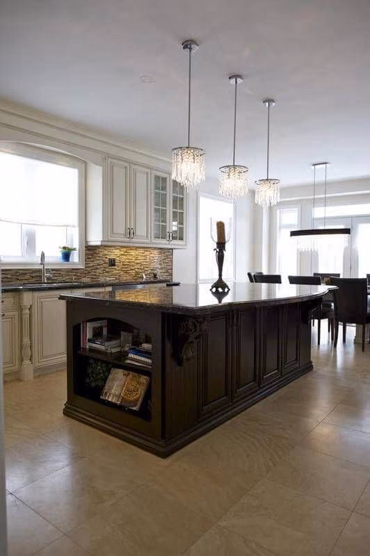 This European-inspired kitchen renovation features a large custom dark wood island with detailed paneling and built-in open shelving, creating both visual impact and functional storage. The contrasting white perimeter cabinetry enhances brightness while maintaining a timeless, classic aesthetic.

The island is topped with premium natural stone, offering ample workspace for cooking and entertaining. Elegant crystal pendant lights above the island add sophistication and warmth, complementing the neutral tile flooring and mosaic backsplash.

This kitchen layout emphasizes spaciousness, symmetry, and refined craftsmanship—ideal for homeowners seeking a high-end European kitchen renovation with a statement island centerpiece.