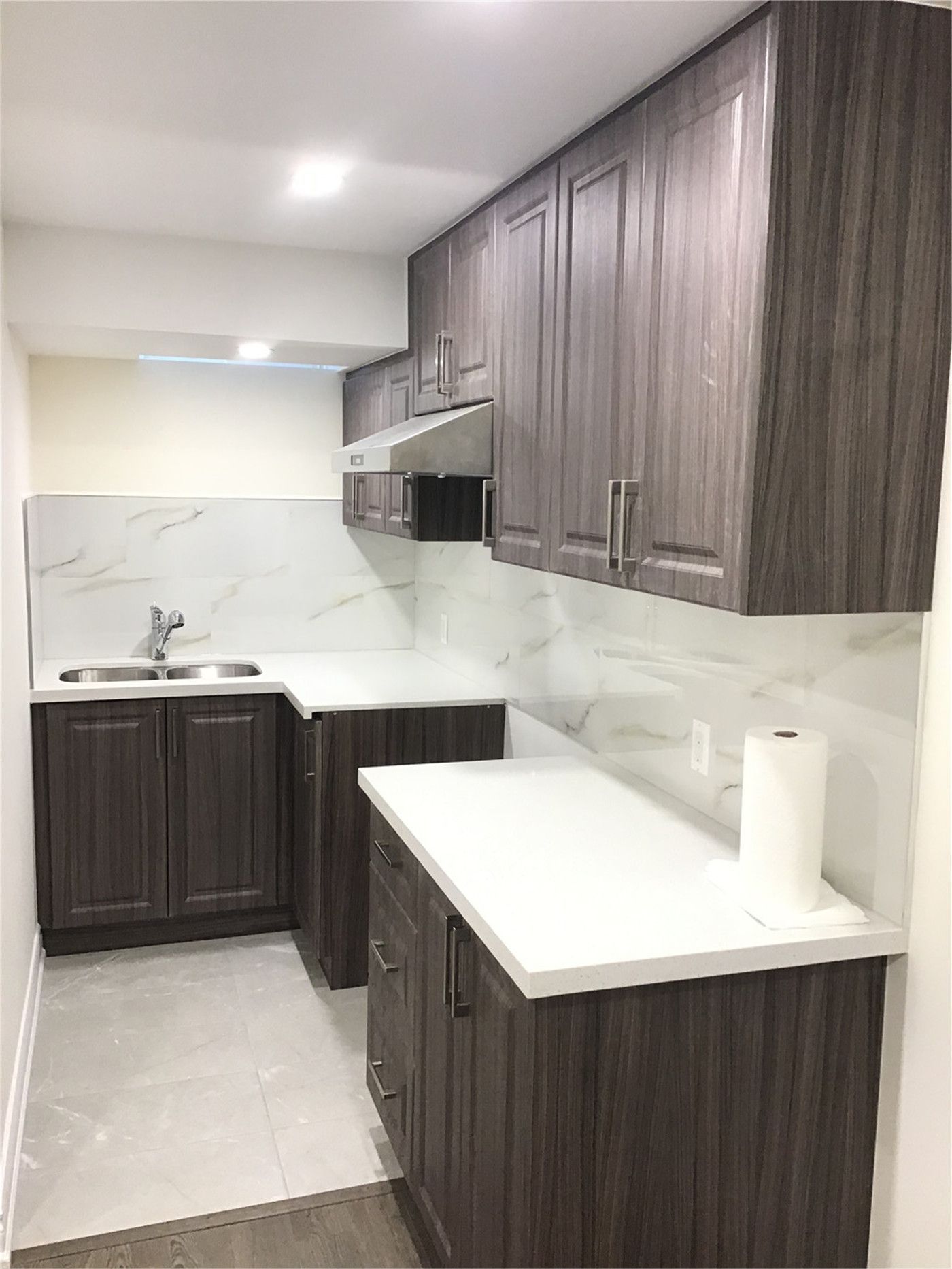 This Mississauga basement renovation includes a compact and modern kitchenette featuring dark wood cabinets, a sleek quartz countertop, and marble-style backsplash. The efficient layout maximizes functionality, making it perfect for a secondary suite, rental unit, or in-law living space.