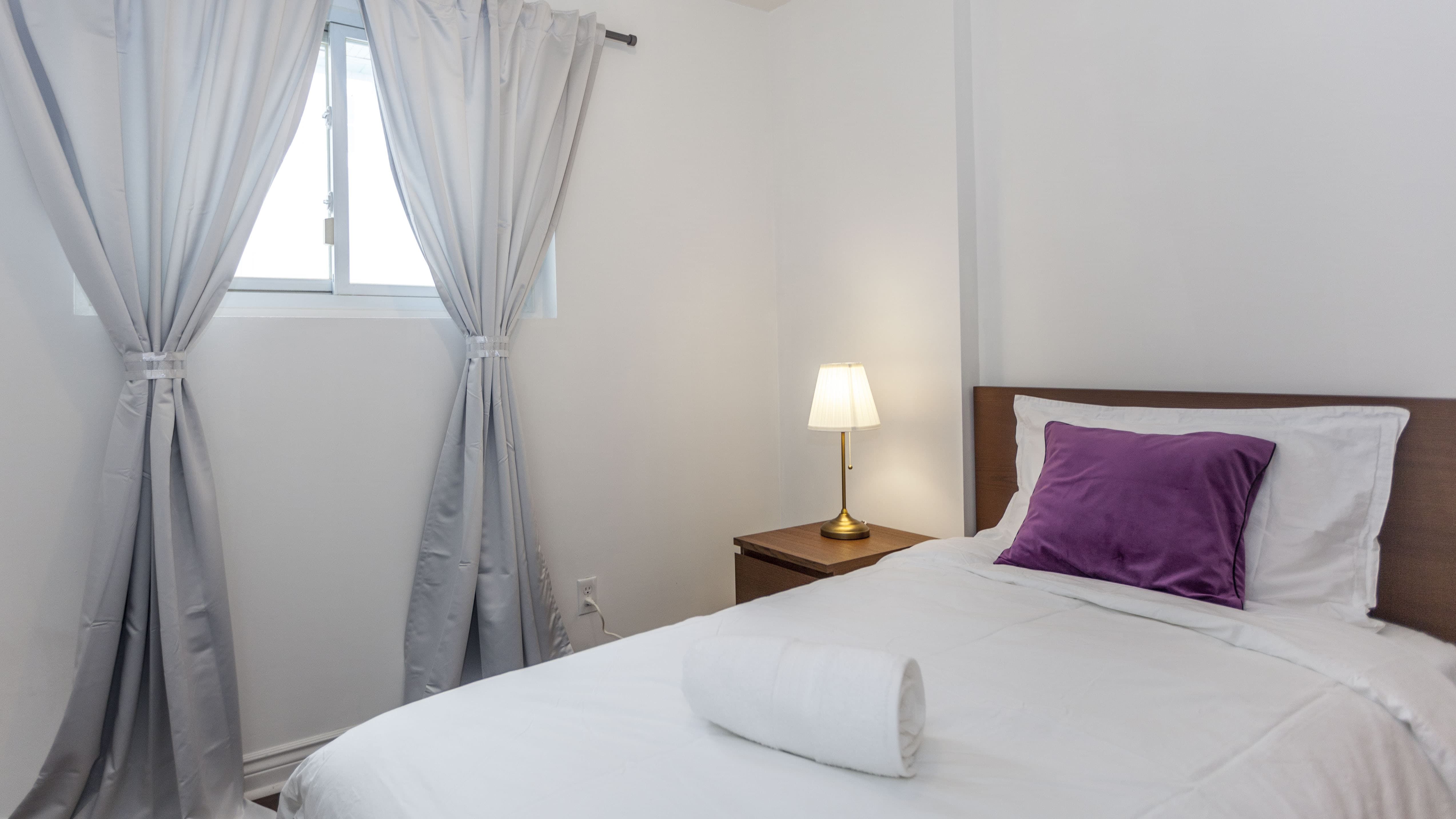 This Toronto bedroom renovation features a bright and airy layout enhanced by natural light from the window and soft curtain design. The clean bedding, wooden nightstand, and warm bedside lighting create a cozy and inviting atmosphere. The space is designed for comfort and relaxation, reflecting the practical and elegant finishing of a professional Toronto Renovation Company.
