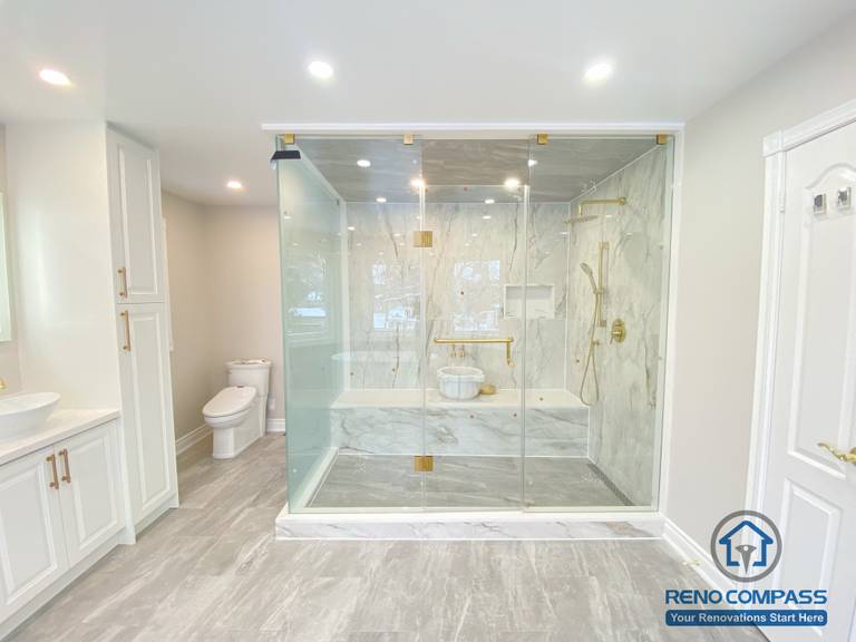 This Richmond Hill bathroom renovation showcases a bright and elegant design featuring a frameless glass shower enclosure, built-in bathtub, marble-style wall tiles, and gold-finish fixtures. The custom white vanity with integrated lighting mirror and large-format floor tiles enhance both functionality and visual openness. This project reflects high-quality craftsmanship and thoughtful layout planning for a modern bathroom renovation in Richmond Hill.