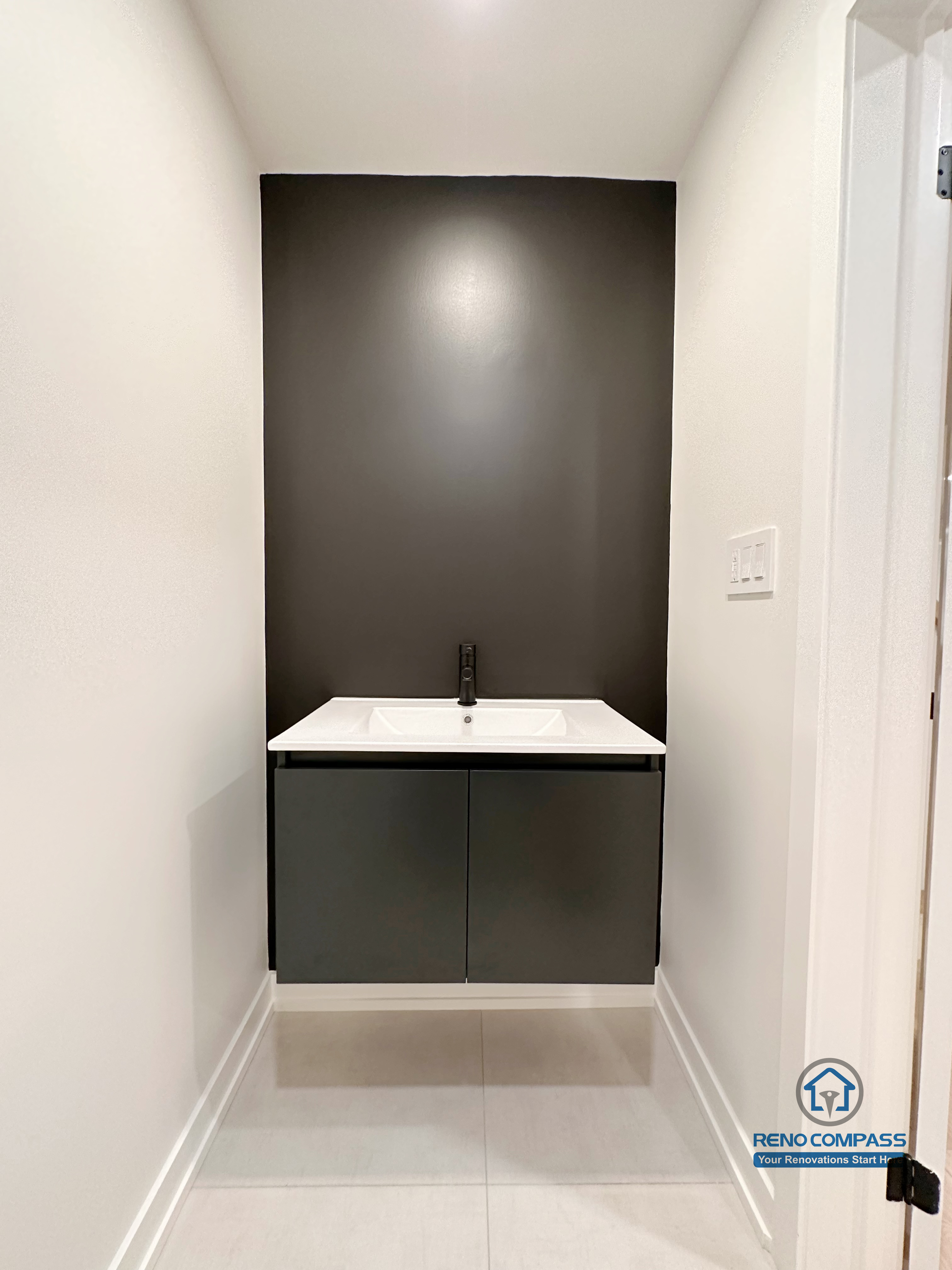 This image features a modern basement powder room completed as part of a Markham basement renovation. The compact vanity design combines a sleek black accent wall, minimalist cabinetry, and a wall-mounted sink to maximize functionality in a limited footprint. Neutral flooring and clean lines create a refined, contemporary look suitable for guest use or entertainment areas. Designed and built by a trusted Toronto renovation company, Reno Compass Inc., this space demonstrates how thoughtful detailing can enhance basement usability and overall home value.