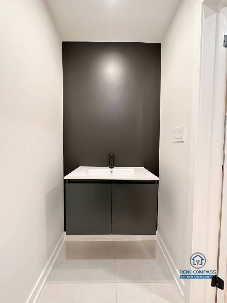 This image features a modern basement powder room completed as part of a Markham basement renovation. The compact vanity design combines a sleek black accent wall, minimalist cabinetry, and a wall-mounted sink to maximize functionality in a limited footprint. Neutral flooring and clean lines create a refined, contemporary look suitable for guest use or entertainment areas. Designed and built by a trusted Toronto renovation company, Reno Compass Inc., this space demonstrates how thoughtful detailing can enhance basement usability and overall home value.