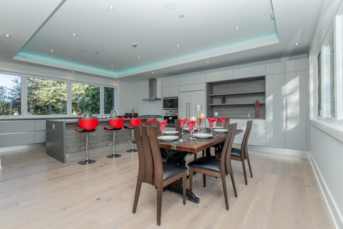 Contemporary open-concept kitchen and dining area in a 5000 sqft Toronto interior renovation, featuring custom grey cabinetry, a large kitchen island with red bar stools, built-in stainless steel appliances, and wide plank hardwood flooring. The dining space is styled with a solid wood table and modern seating, creating a seamless flow between cooking and entertaining areas. Completed by Reno Compass Inc., a trusted Toronto renovation company specializing in luxury home interior remodeling and large-scale residential upgrades.