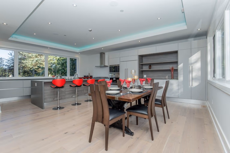 Contemporary open-concept kitchen and dining area in a 5000 sqft Toronto interior renovation, featuring custom grey cabinetry, a large kitchen island with red bar stools, built-in stainless steel appliances, and wide plank hardwood flooring. The dining space is styled with a solid wood table and modern seating, creating a seamless flow between cooking and entertaining areas. Completed by Reno Compass Inc., a trusted Toronto renovation company specializing in luxury home interior remodeling and large-scale residential upgrades.