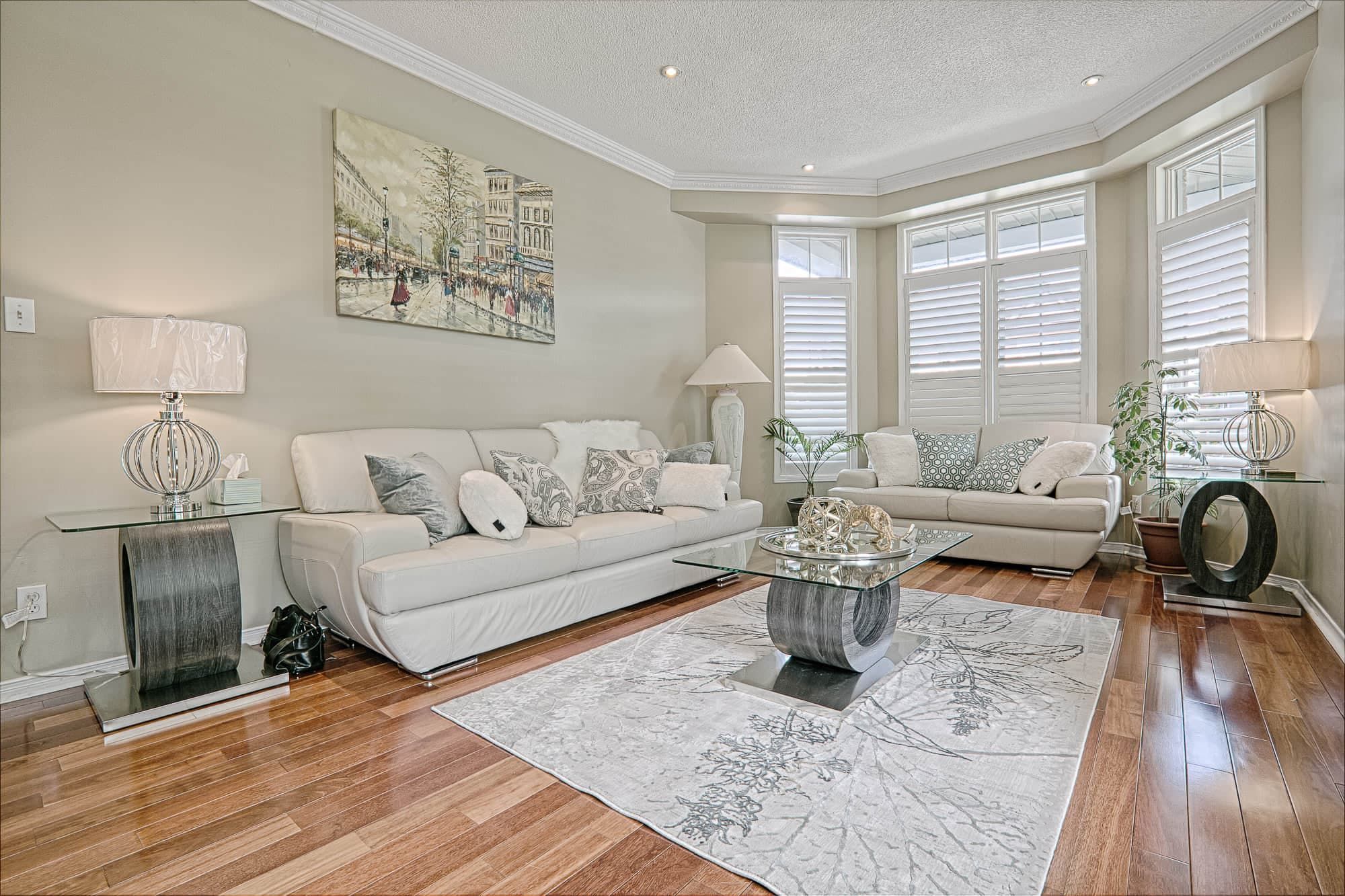 This elegant living room interior showcases warm hardwood flooring, soft neutral wall tones, and coordinated modern furniture that create a bright and sophisticated atmosphere. The large bay windows with plantation shutters allow abundant natural light, while the contemporary coffee table, textured area rug, and layered lighting enhance comfort and style. A refined example of warm minimalist living room renovation and modern home interior design inspiration for homeowners in Toronto and the GTA.