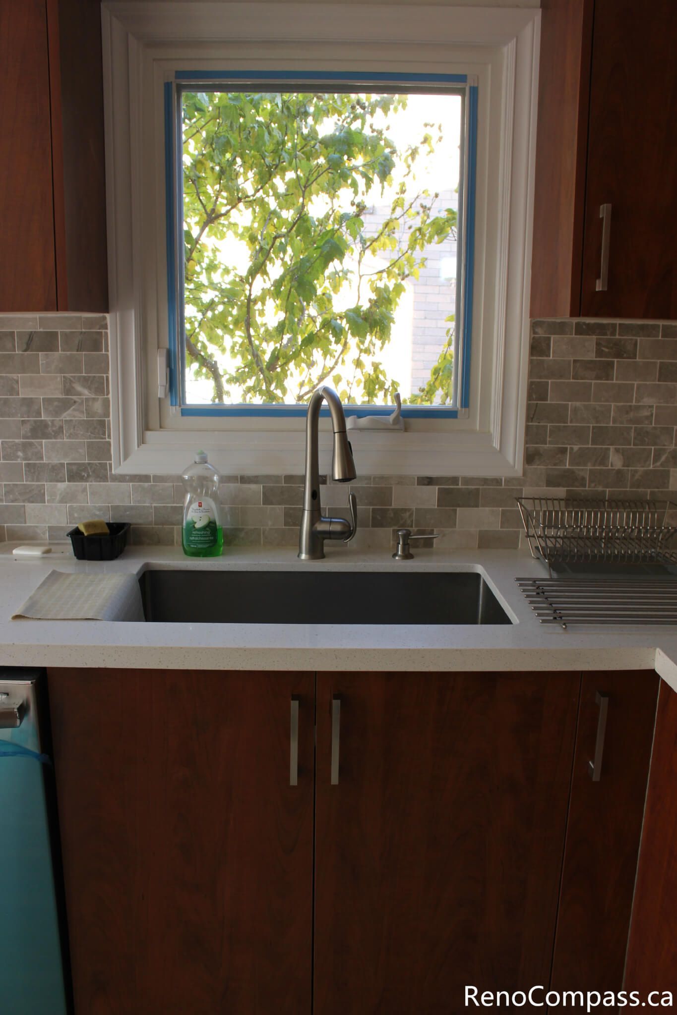 This bright kitchen sink area is part of a full home renovation project in Mississauga completed by a professional Toronto renovation company. The design features a modern undermount sink, sleek faucet, quartz countertop, and neutral-toned mosaic backsplash, complemented by natural light from the window. The renovation enhances everyday functionality, lighting, and visual comfort while creating a clean, modern kitchen space that blends durability with contemporary style.