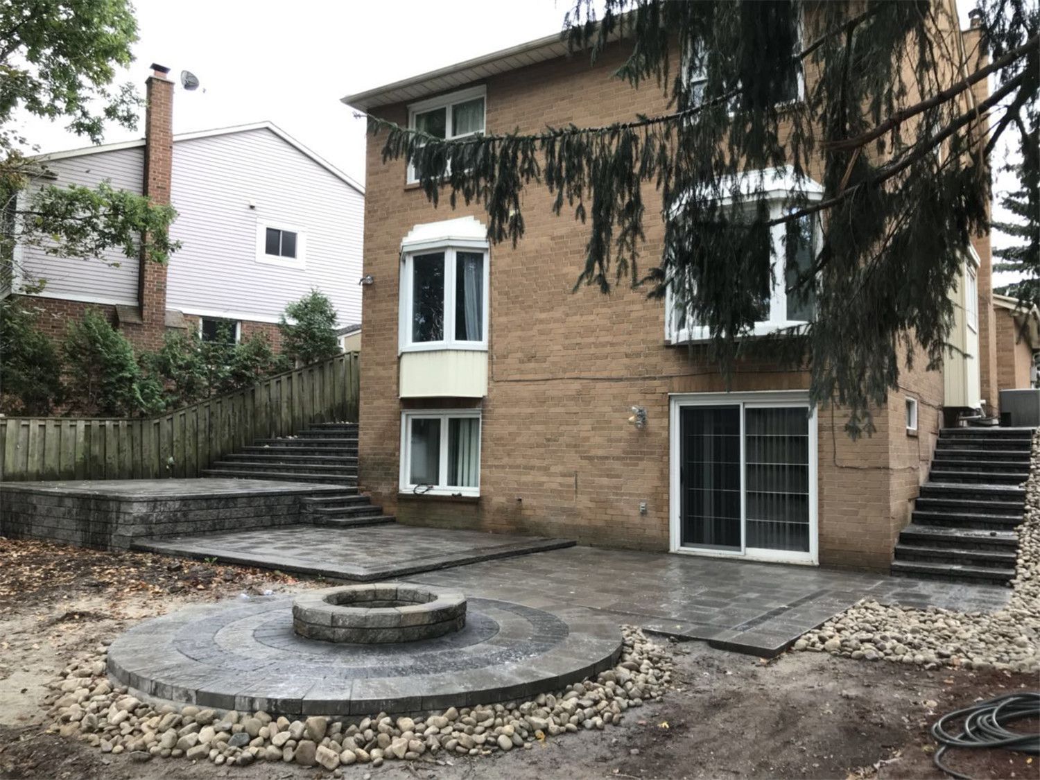 Backyard landscaping project in Toronto featuring a circular stone fire pit patio, natural rock border, and multi-level terrace steps. The design creates a relaxing outdoor gathering space while improving the backyard layout and functionality.