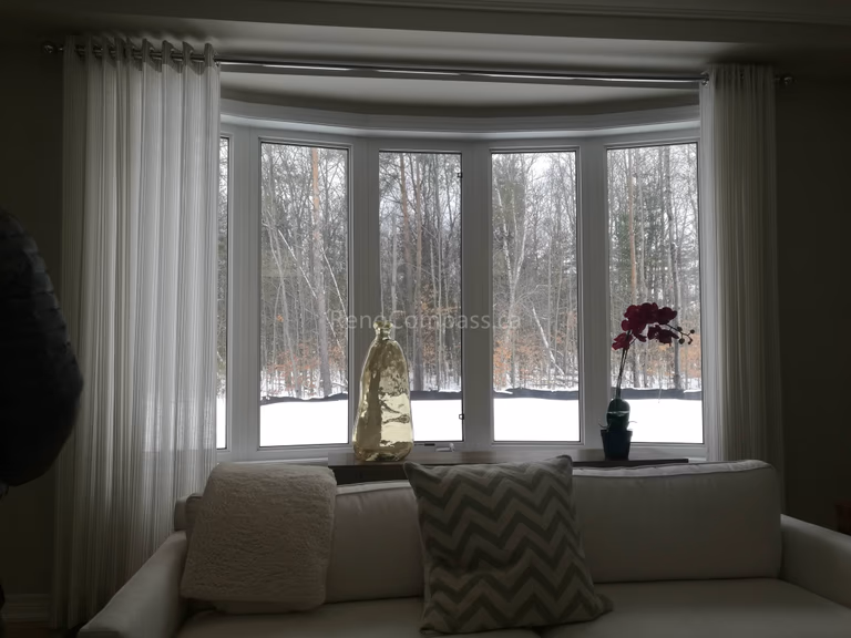 This image showcases a beautifully finished living room as part of a Toronto interior renovation project. The curved bay window design allows abundant natural light to flow into the space, creating a calm and inviting atmosphere. Soft white curtains, clean window framing, and a neutral sofa setup enhance the overall brightness and comfort of the room. The renovation emphasizes natural views, elegant proportions, and a timeless interior aesthetic, reflecting high-quality interior renovation craftsmanship in Toronto homes.