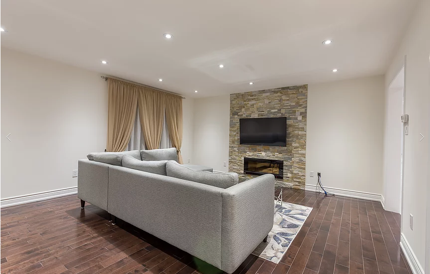 A welcoming modern living room from our Vaughan full house renovation featuring a stone fireplace wall, mounted TV, and elegant neutral décor. The comfortable sectional sofa and hardwood flooring create a perfect balance of style and warmth for everyday family living.