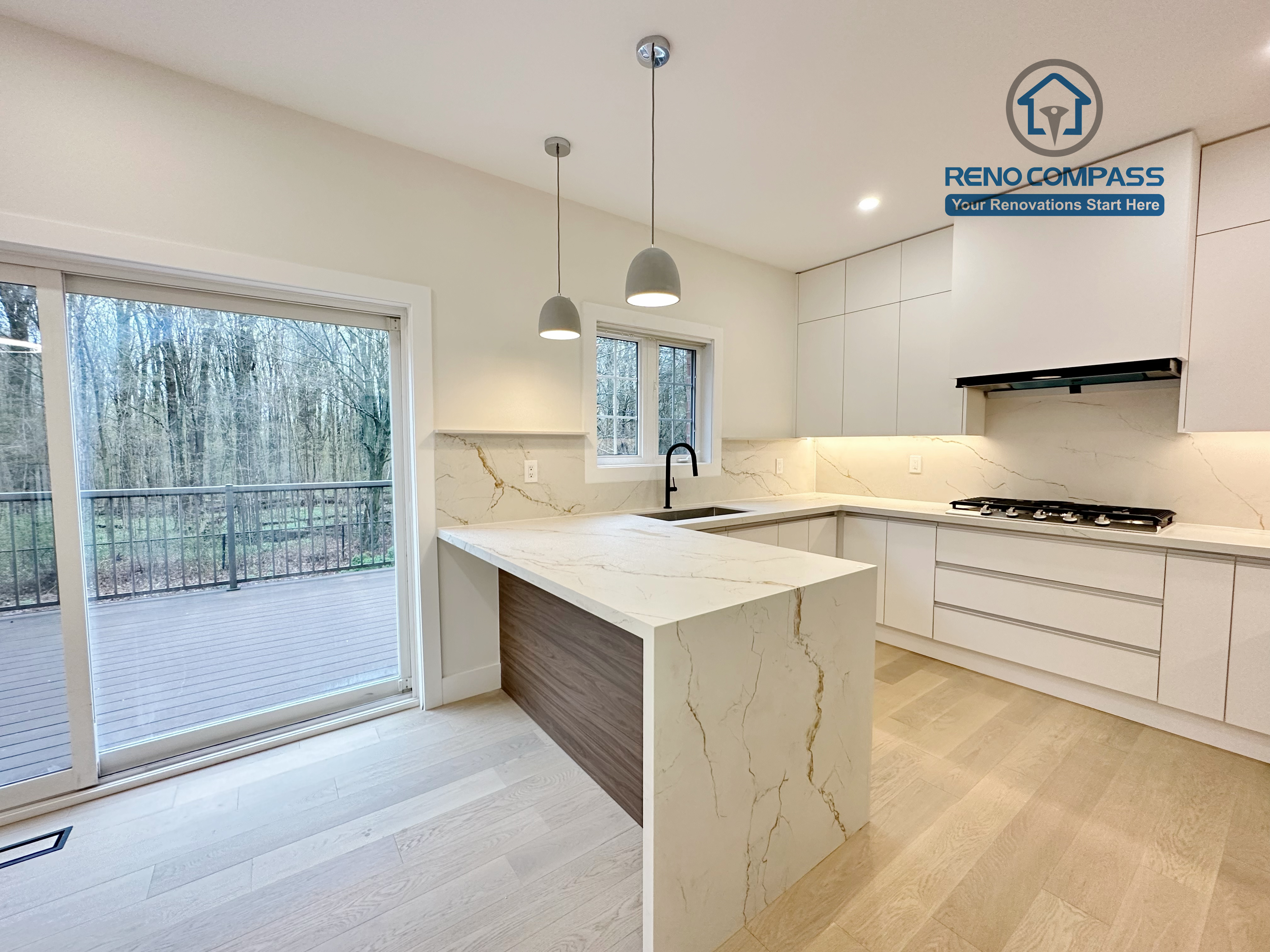 This image features a modern kitchen renovation in Richmond Hill with a quartz waterfall island showcasing natural veining and warm wood panel accents. The open-concept layout includes custom white cabinetry, a built-in gas cooktop, minimalist pendant lighting, and seamless access to the outdoor patio through large sliding doors. Natural light and light-tone hardwood flooring enhance the clean, airy atmosphere. Completed by a trusted Toronto Renovation Company, this kitchen renovation balances elegant design, functional workflow, and indoor-outdoor living.