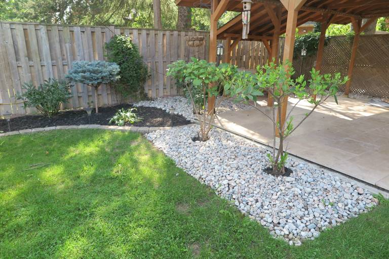 This image features a completed North York backyard landscaping renovation combining a wooden pergola, decorative stone garden beds, and newly planted shrubs. The layout improves drainage, defines outdoor zones, and creates a clean transition between lawn, garden, and patio areas. Designed and built by a Toronto Renovation Company specializing in residential landscaping and outdoor living spaces.