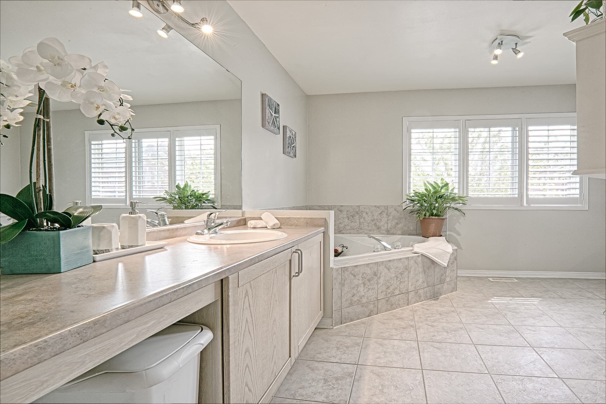 This warm minimalist bathroom design features a spacious built-in soaking tub, large vanity with extended countertop, and soft neutral tile finishes that create a relaxing and elegant atmosphere. The oversized mirror enhances natural light, while subtle décor elements and indoor plants add warmth and balance to the space. A perfect example of modern, cozy bathroom interior design for homeowners seeking functional yet stylish renovation ideas in Toronto and the GTA.