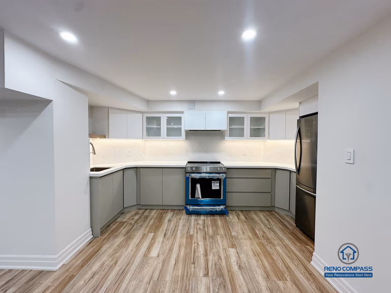 This image shows a completed basement kitchen renovation in Pickering, captured from a front-facing perspective to highlight the symmetrical layout and clean cabinet alignment. Custom cabinetry, integrated under-cabinet lighting, and a continuous countertop surface create a functional and visually balanced workspace. Neutral tones and consistent flooring help the kitchen blend seamlessly into the surrounding basement living area, supporting everyday use without visual clutter. Recessed ceiling lighting enhances brightness and clarity, reinforcing a practical, well-organized lower-level kitchen design.
