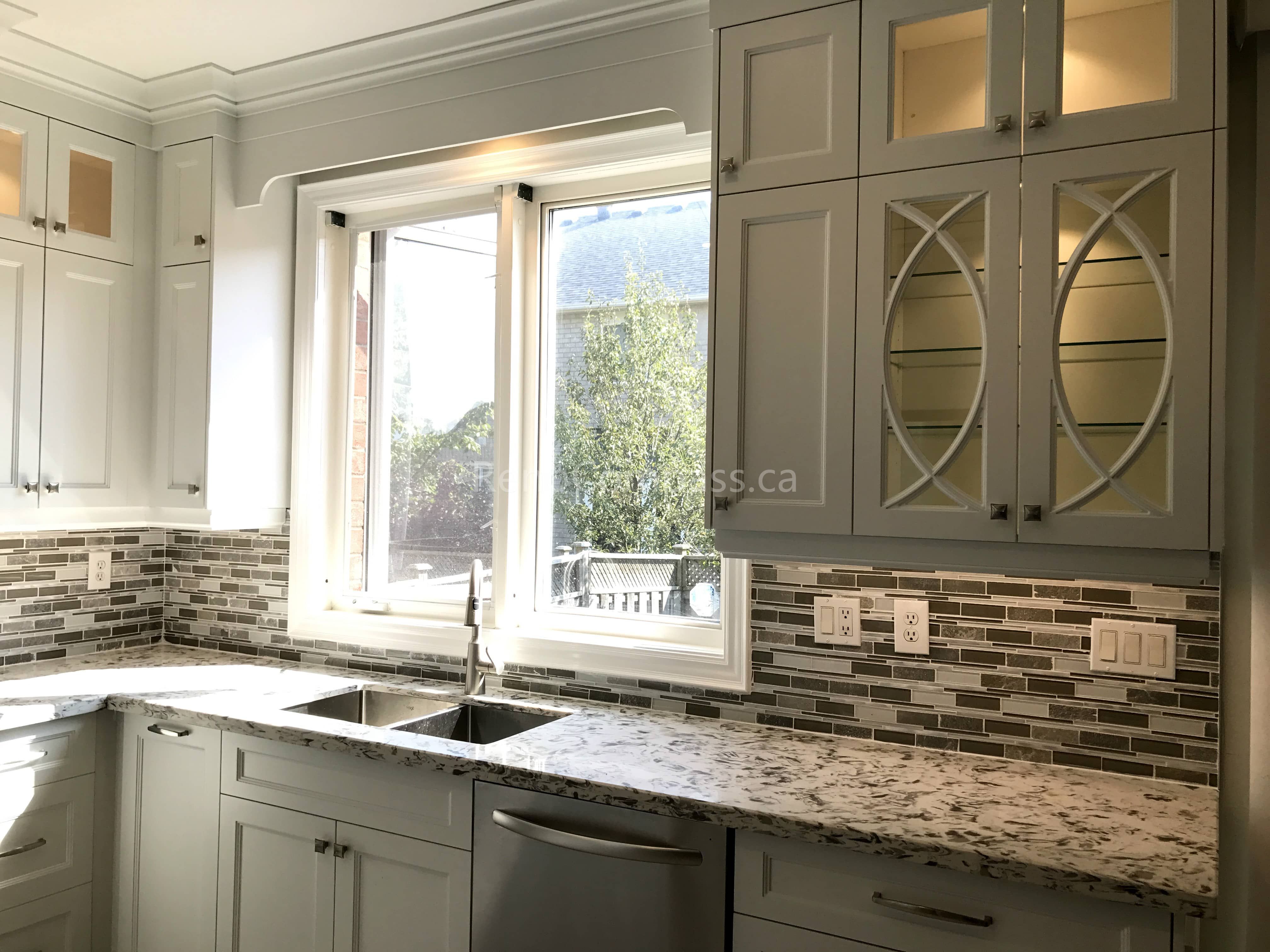 This Markham kitchen renovation highlights a bright double-sink workstation under a large window, paired with custom cabinetry and granite countertops. Decorative glass cabinet inserts and modern mosaic backsplash tiles add elegance, while the updated layout enhances natural light, storage, and daily functionality for a refined modern kitchen space.