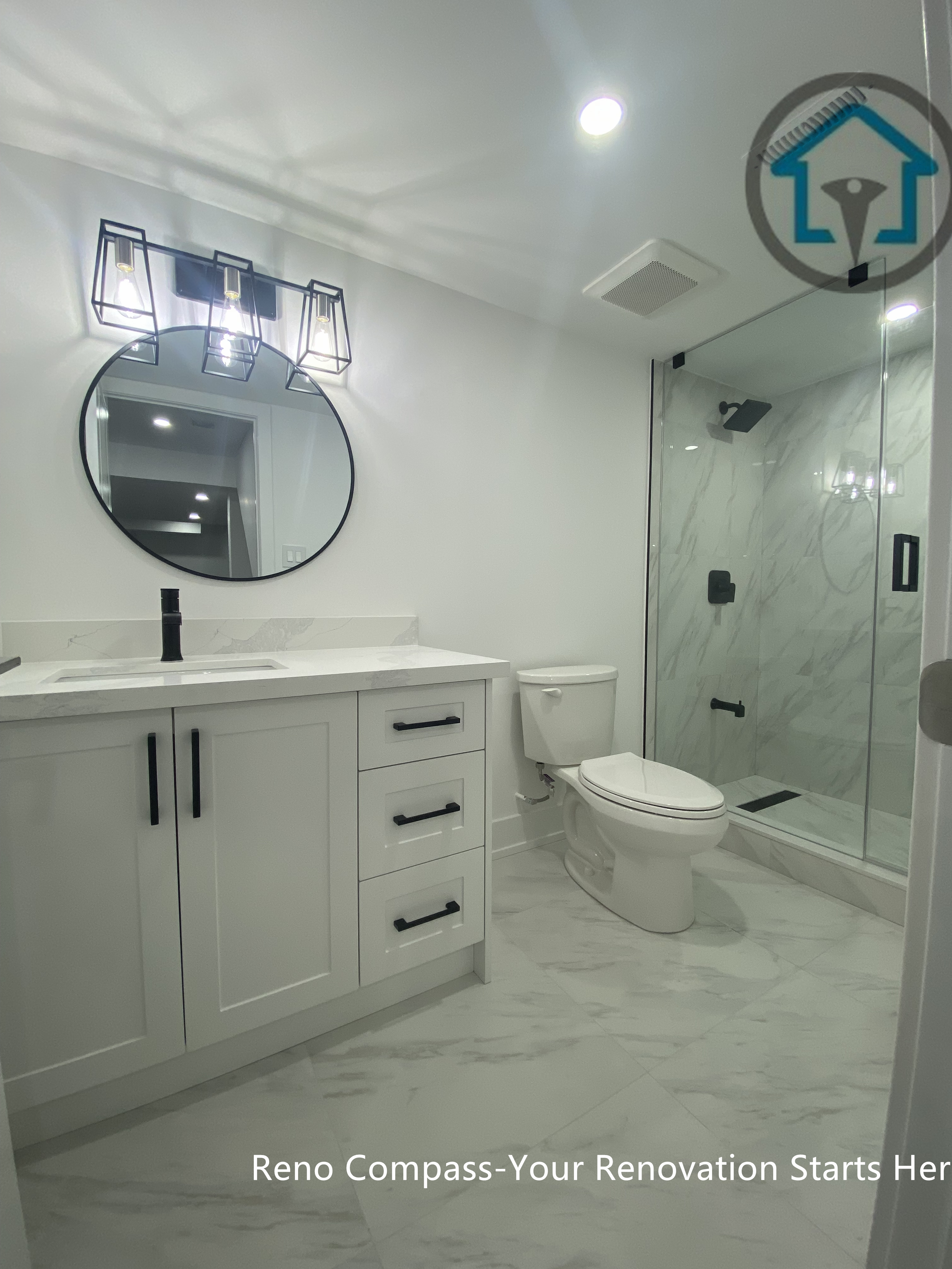 This image features a full basement bathroom renovation in Newmarket completed by a professional Toronto renovation company. The space includes a custom white vanity with quartz countertop, modern black fixtures, a round mirror with contemporary wall lighting, and a frameless glass walk-in shower finished with marble-look tiles. The clean layout and durable materials create a bright, functional, and stylish basement bathroom suitable for long-term family use or rental properties.