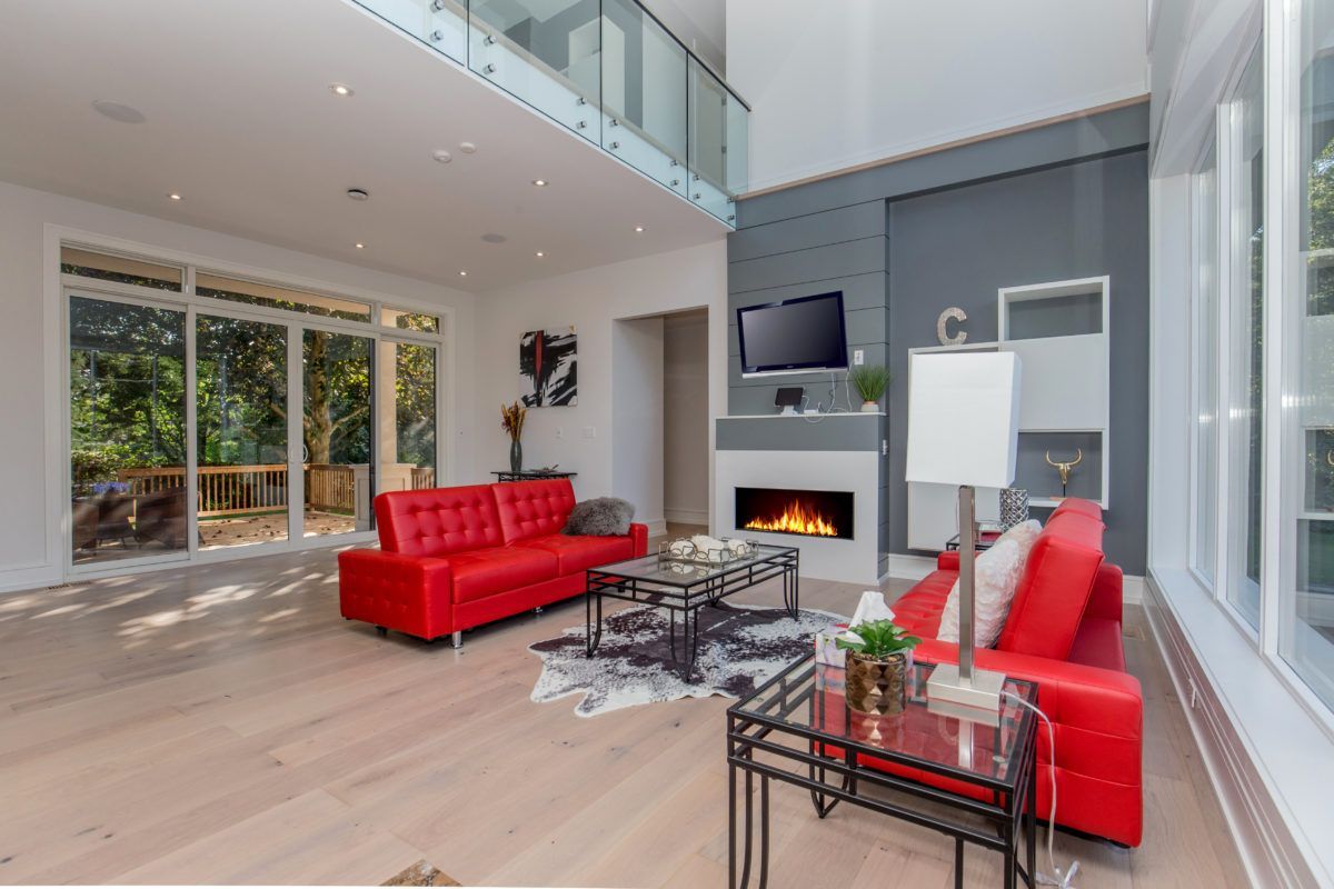 Modern open-concept living room in a 5000 sqft Toronto home renovation featuring double-height ceilings, glass railing mezzanine, wide plank hardwood flooring, contemporary fireplace wall with built-in TV, and large sliding doors leading to a backyard deck. The space combines natural light, clean architectural lines, and bold red leather sofas to create a bright and sophisticated interior. Renovated by Reno Compass Inc., a professional Toronto renovation company specializing in large-scale custom home interior transformations.