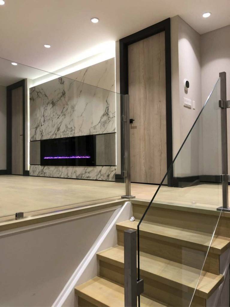 Modern basement renovation in Markham showcasing a sleek marble-style feature wall with built-in electric fireplace, recessed pot lights, and wide plank hardwood flooring. The open-concept layout includes glass railing stairs and contemporary wood doors, creating a bright, inviting entertainment space designed for comfort, style, and added home value.