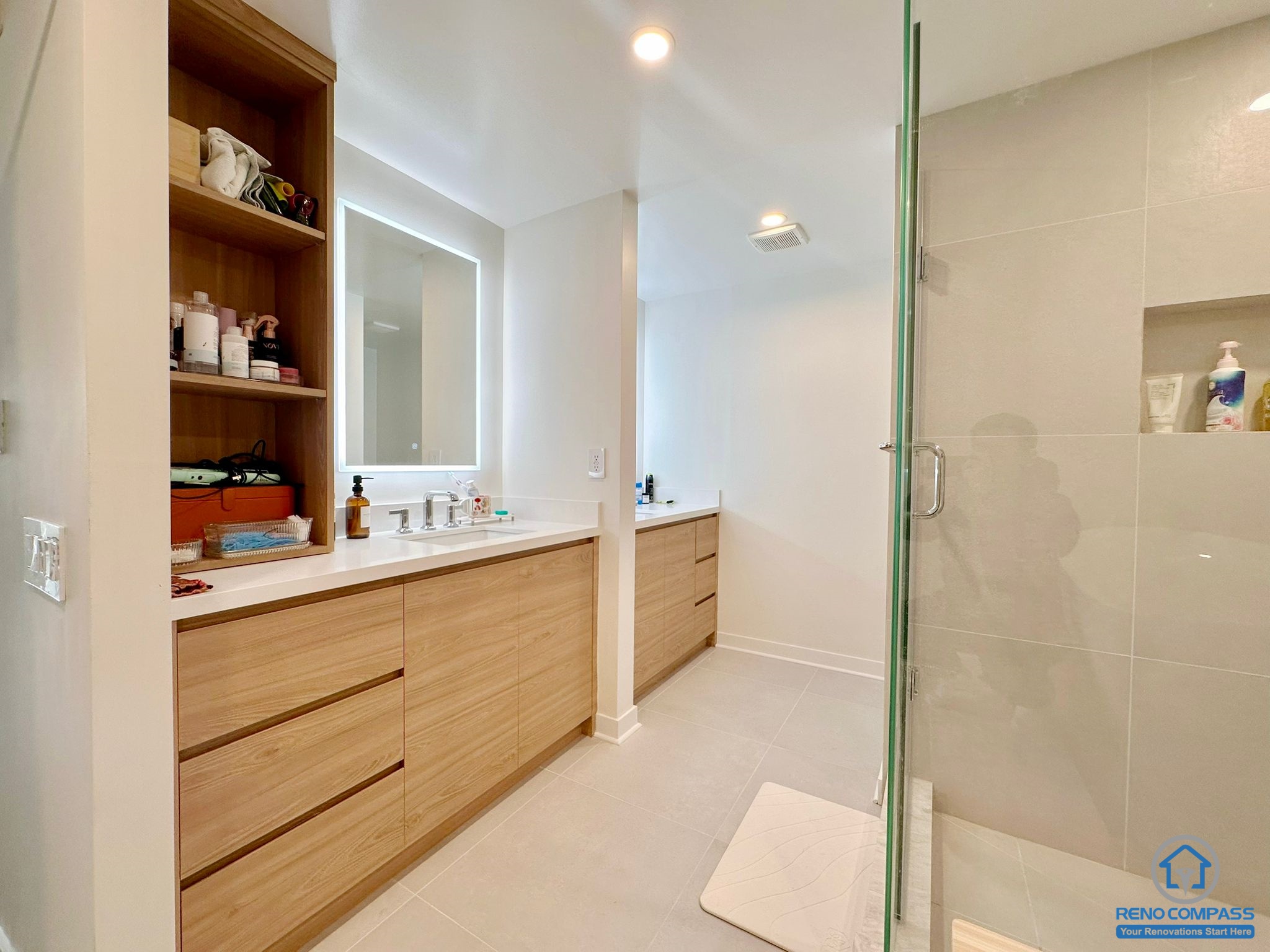 This Toronto condo master bathroom renovation features a stylish double vanity design separated by a partial wall for added privacy and functionality. Both sides are equipped with modern LED mirrors, creating a bright and elegant atmosphere. Custom wood cabinetry offers extensive storage space, while the sleek glass shower enclosure at the back enhances the contemporary and spacious feel of this luxurious bathroom.