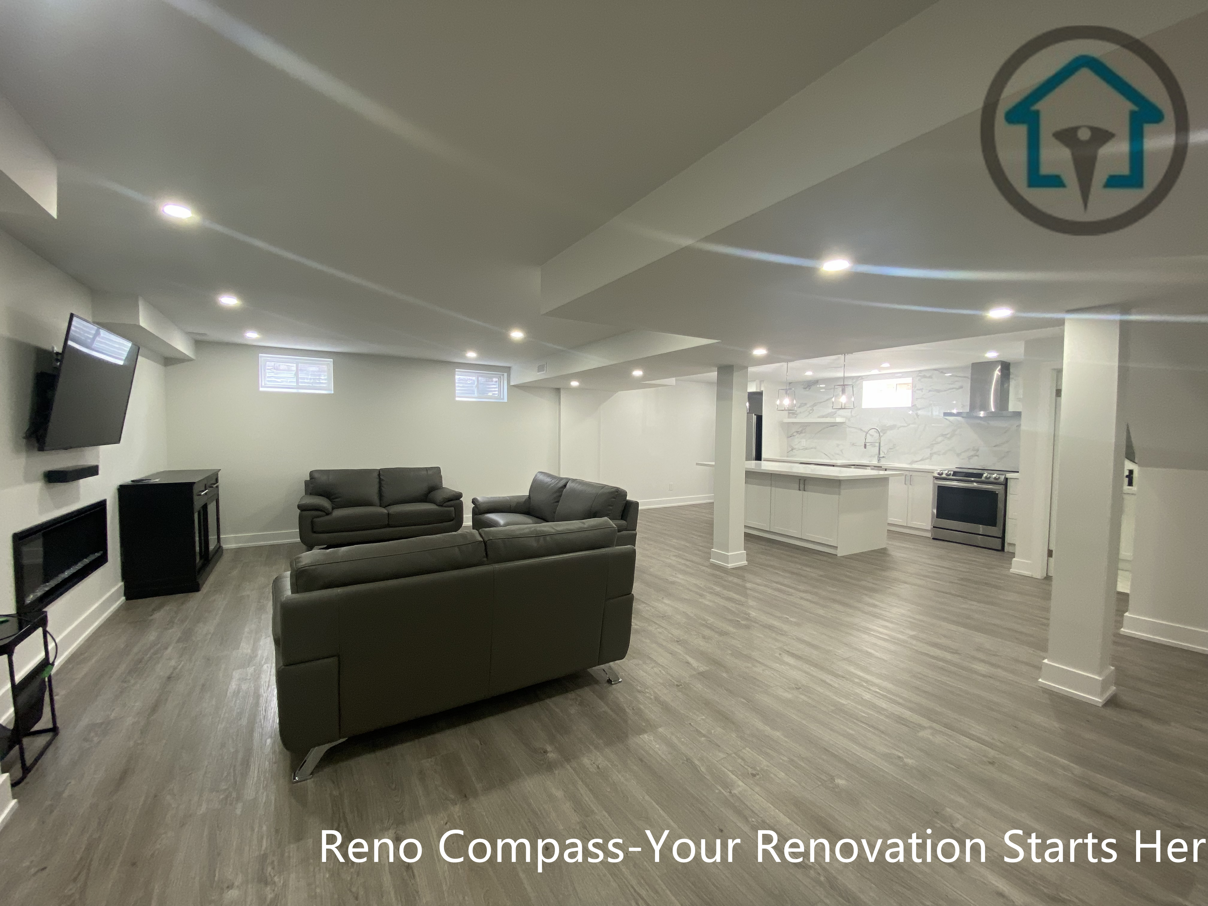 This image presents a spacious open-concept basement renovation in Newmarket completed by a professional Toronto renovation company. The layout integrates a large family living area with modern recessed lighting, wood-look flooring, and structural columns that define zones without closing off the space. The open kitchen with a central island is visible in the background, creating a cohesive and flexible basement layout ideal for entertainment, family gatherings, or rental use.