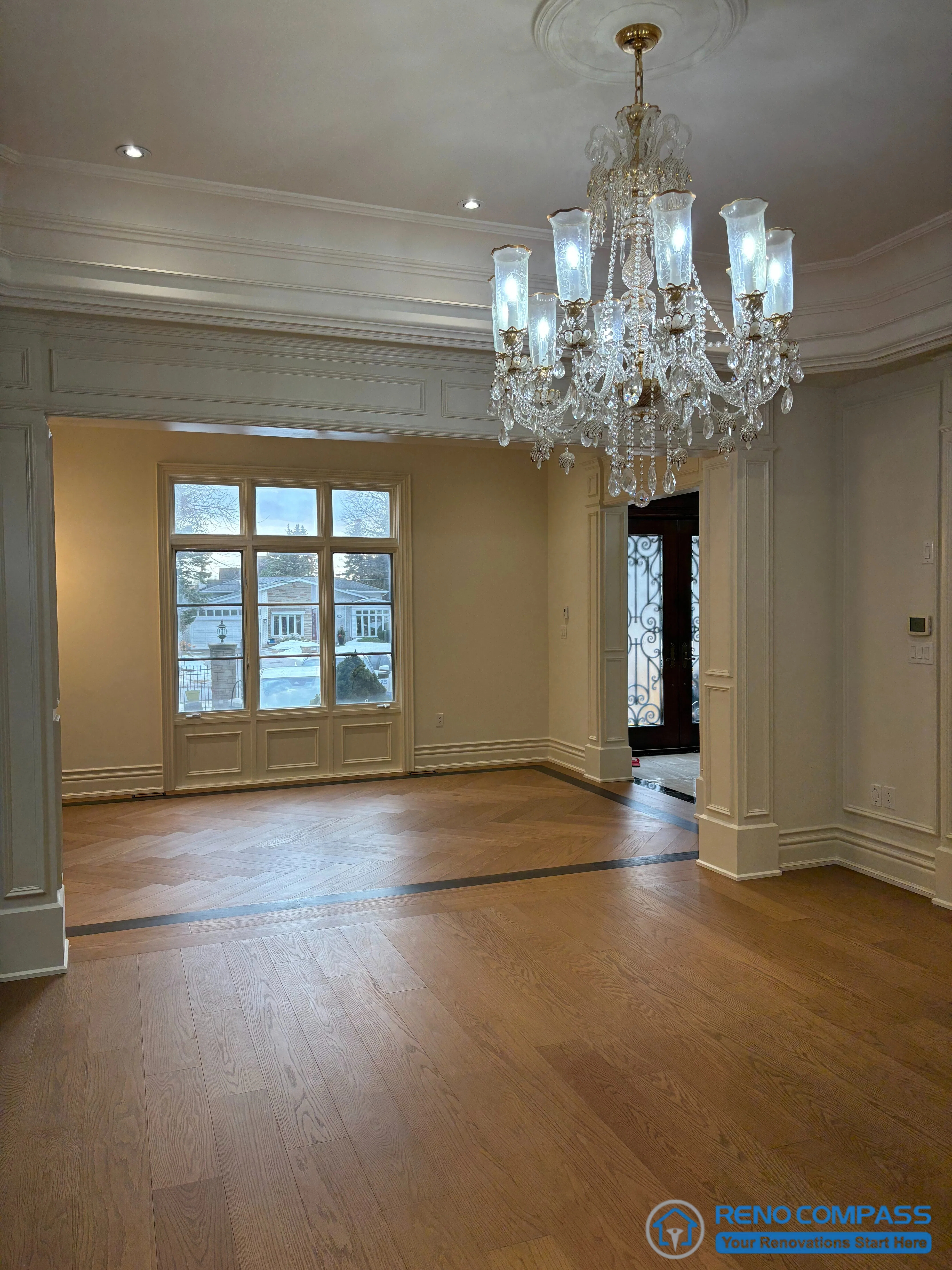 This image presents a European-style living space defined by a centrally positioned crystal chandelier, layered ceiling moulding, and clearly articulated architectural openings. The chandelier establishes a formal vertical axis, while the surrounding ceiling details reinforce proportion and symmetry. Hardwood flooring anchors the room and guides movement toward adjacent spaces, maintaining architectural continuity throughout the interior. Wall panelling and trim are treated as structural elements rather than surface decoration, reflecting a composed and disciplined approach to European interior design within a high-end Richmond Hill interior renovation.