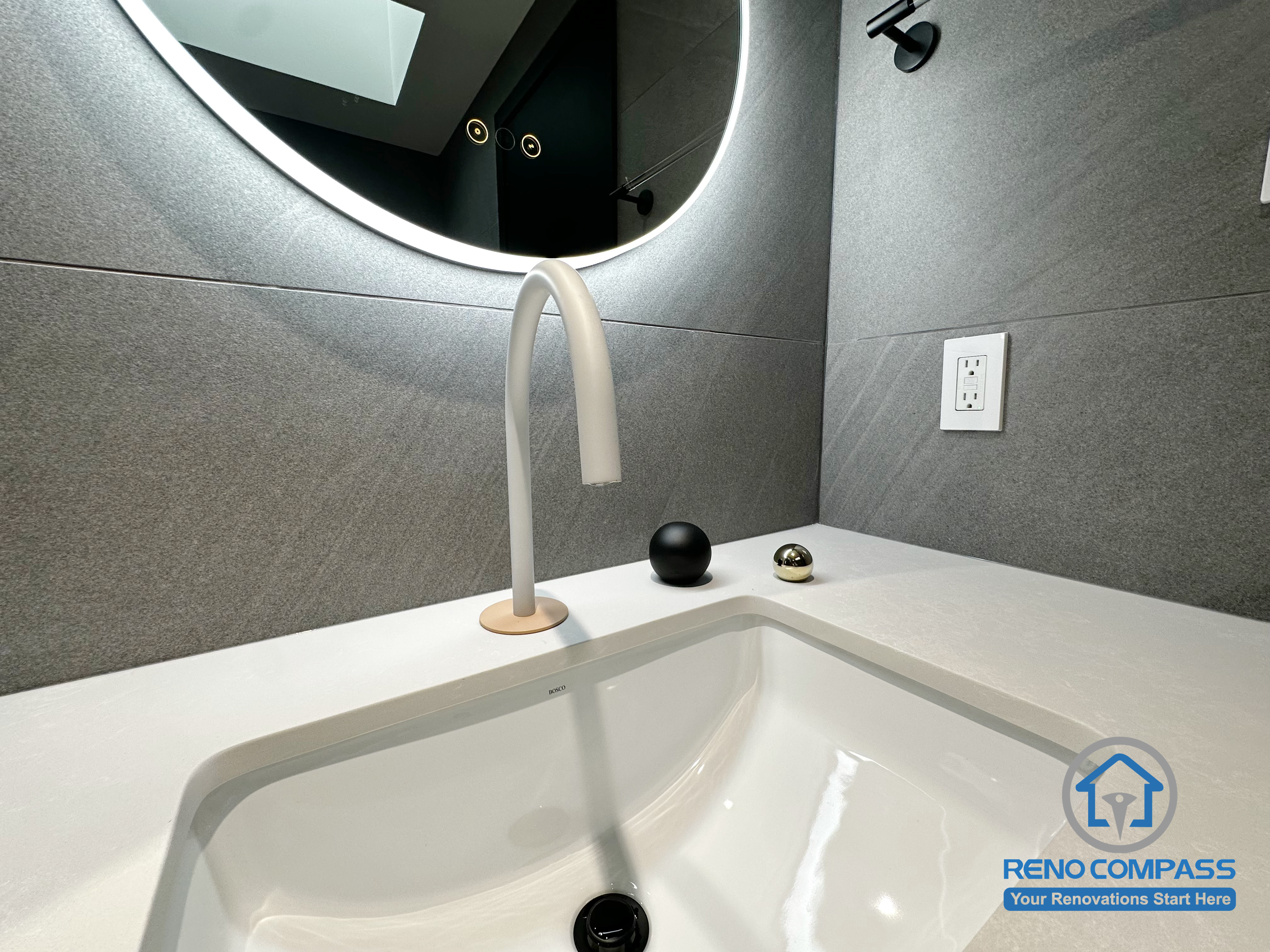 A close-up view of the custom bathroom vanity in this Richmond Hill renovation, featuring a sleek integrated sink, a sculptural modern faucet, and soft LED backlit mirror. The neutral grey tile backdrop and minimalist accessories highlight clean lines and refined material choices, creating a calm and contemporary bathroom atmosphere. Designed and completed by Reno Compass for a modern GTA home.